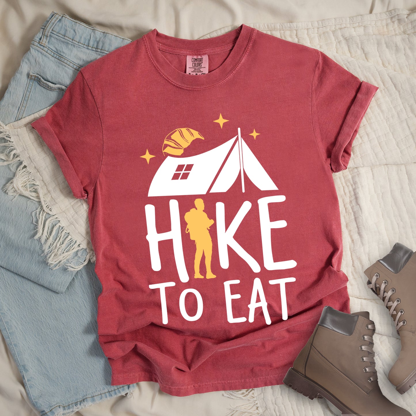 Crimson (Dark Pink) graphic t-shirt with 'Hike to Eat' text featuring white camping tent, yellow hiker silhouette, croissant, and stars design, styled with light wash jeans and brown hiking boots.