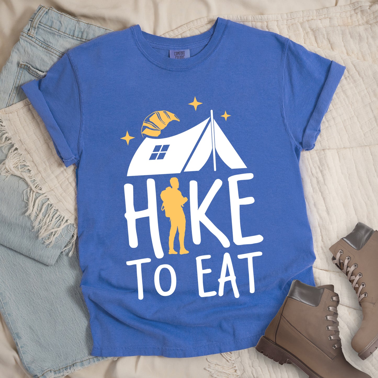 Flo Blue (Bright Blue) graphic t-shirt with 'Hike to Eat' text featuring white camping tent, yellow hiker silhouette, croissant, and stars design, styled with light wash jeans and brown hiking boots.