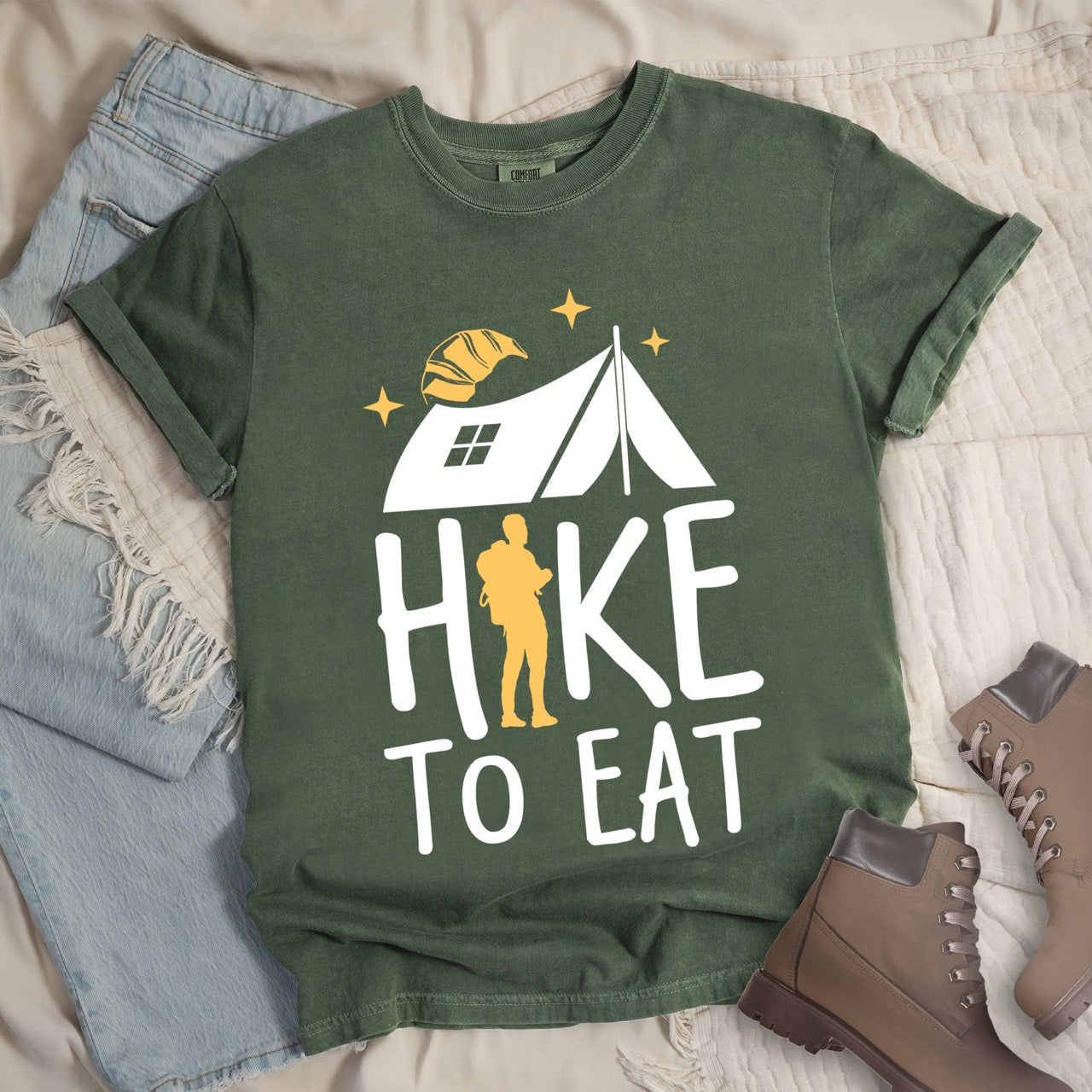 Moss (green) Hike to Eat brand philosophy vintage tee with white camping tent croissant moon stars character silhouette with backpack integrated into typography on Comfort Colors shirt for food motivated adventurers, outdoor enthusiasts and hikers - laid flat with boots and jeans
