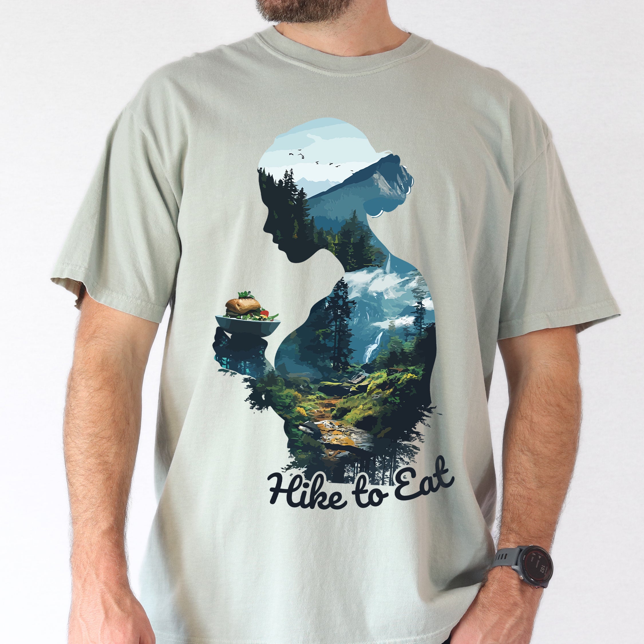 A man wearing a Wilderness Within Hike to Eat t-shirt featuring double exposure design of hiker silhouette holding plate with sandwich / burger merged with mountain forest landscape on Comfort Colors shirt in bay color.