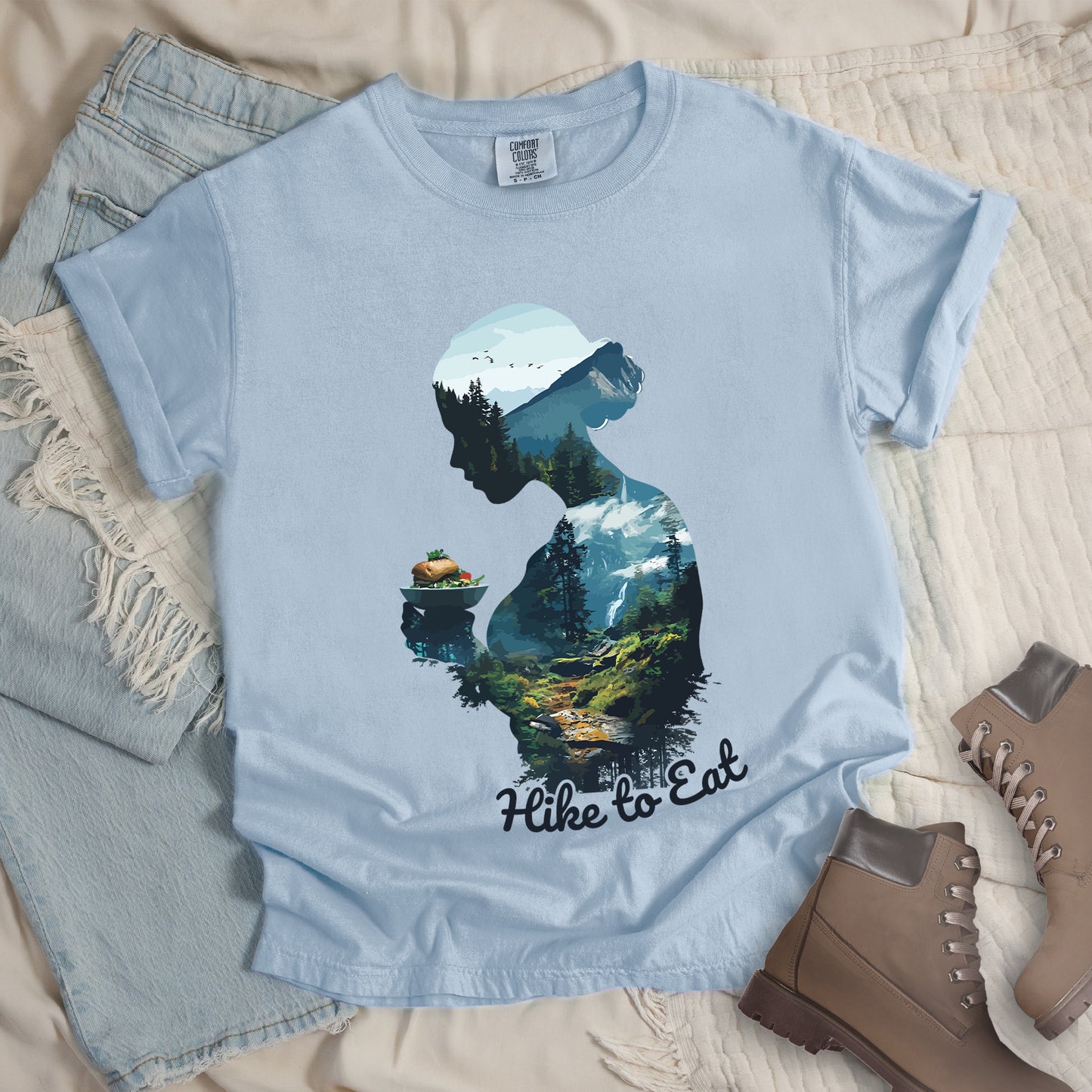 Chambray (Light Blue) graphic t-shirt with 'Hike to Eat' text and 'Wilderness Within' design featuring woman's silhouette filled with mountain landscape, pine trees, and hiking trail, styled with light wash jeans and brown hiking boots