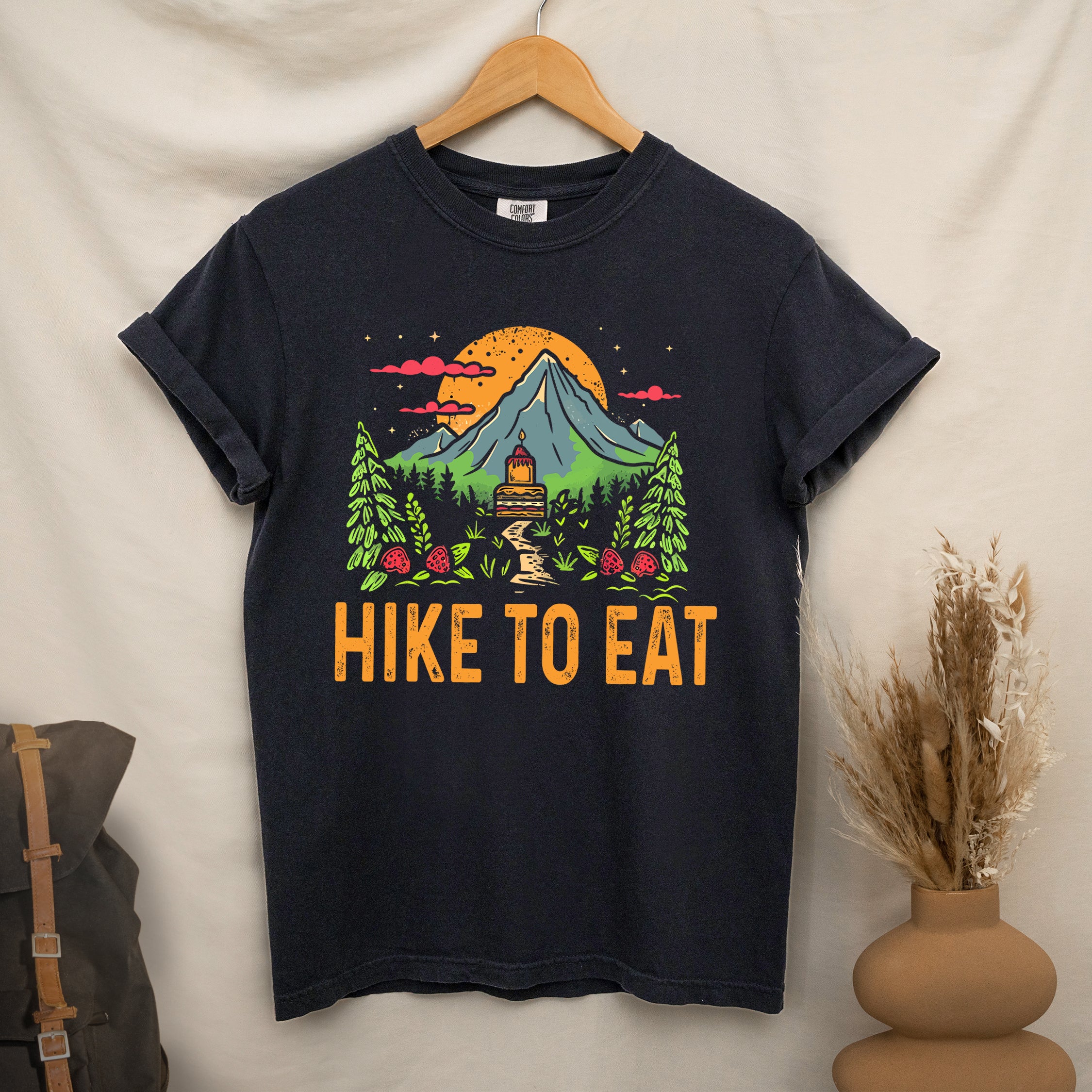 Black graphic t-shirt on a wooden hanger with 'Hike to Eat' text and 'Mountain Sweets' design featuring layered cake in mountain landscape with orange sunset, pine trees, mushrooms, and starry sky