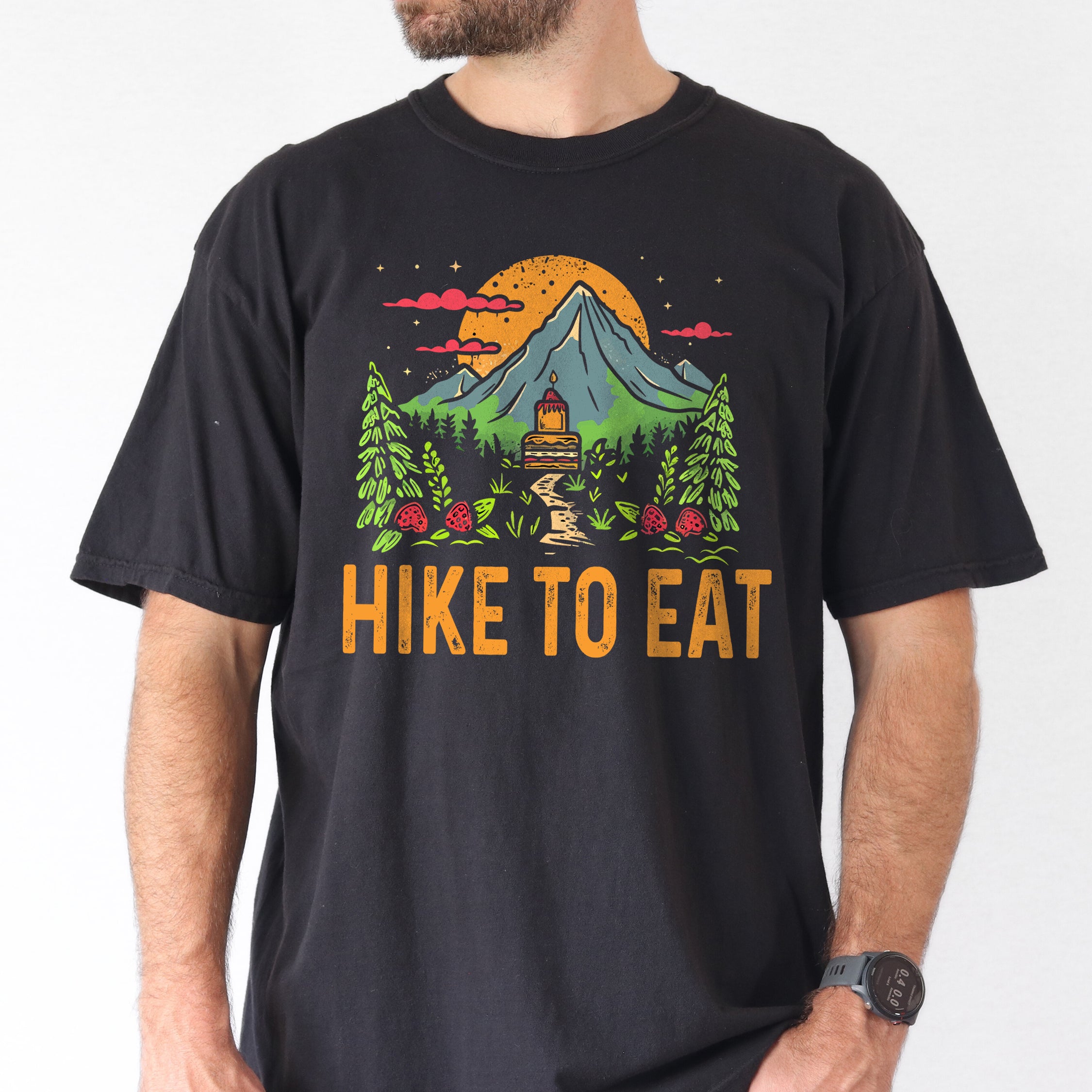Black graphic t-shirt with 'Hike to Eat' text and 'Mountain Sweets' design featuring layered cake in mountain landscape with orange sunset, pine trees, mushrooms, and starry sky worn by a man.