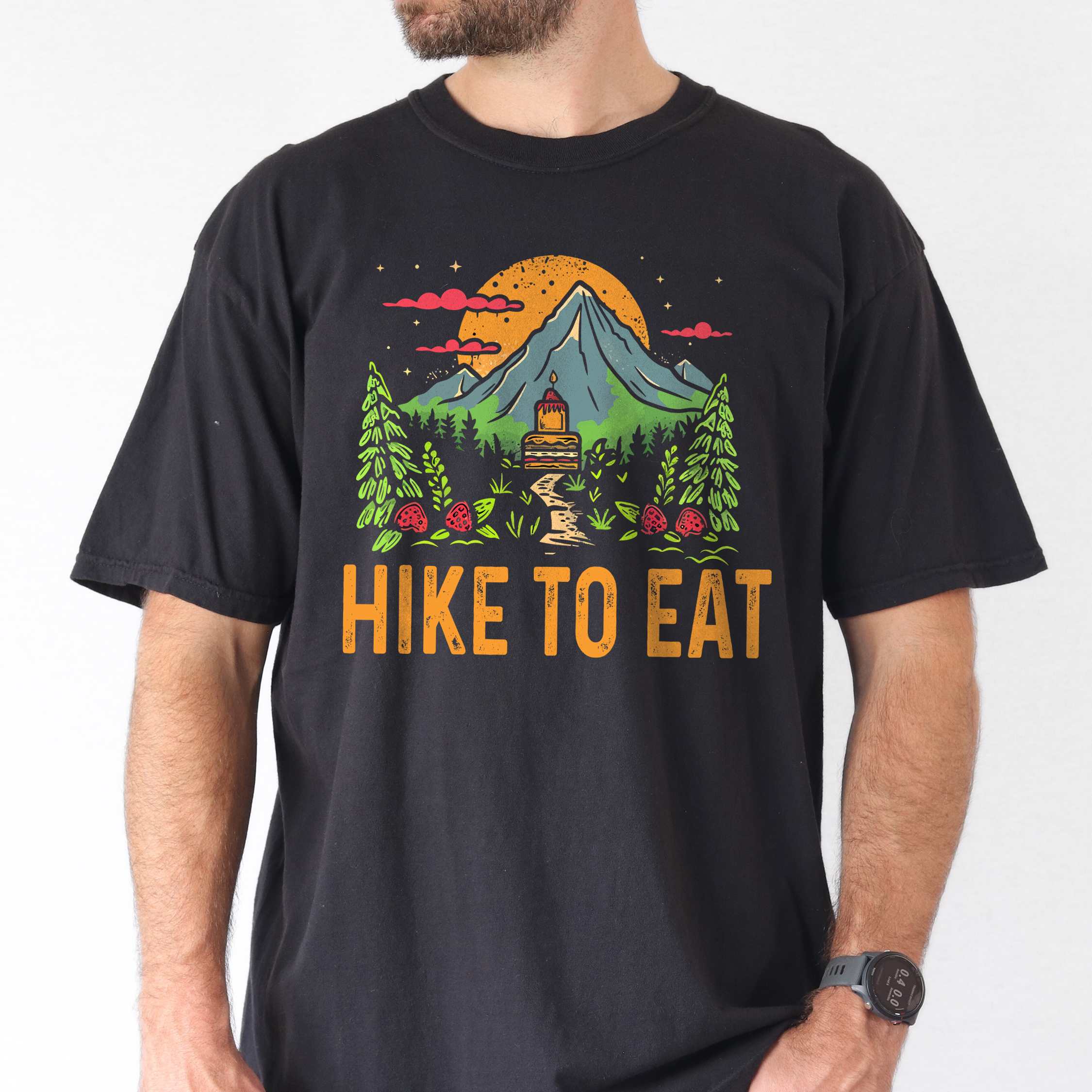 Man wearing black "Hike to Eat" colored vintage statement tee with text in cream lettering, mountains, birds, silhouette, illustrated tiered cake, and perfect for cake lovers.