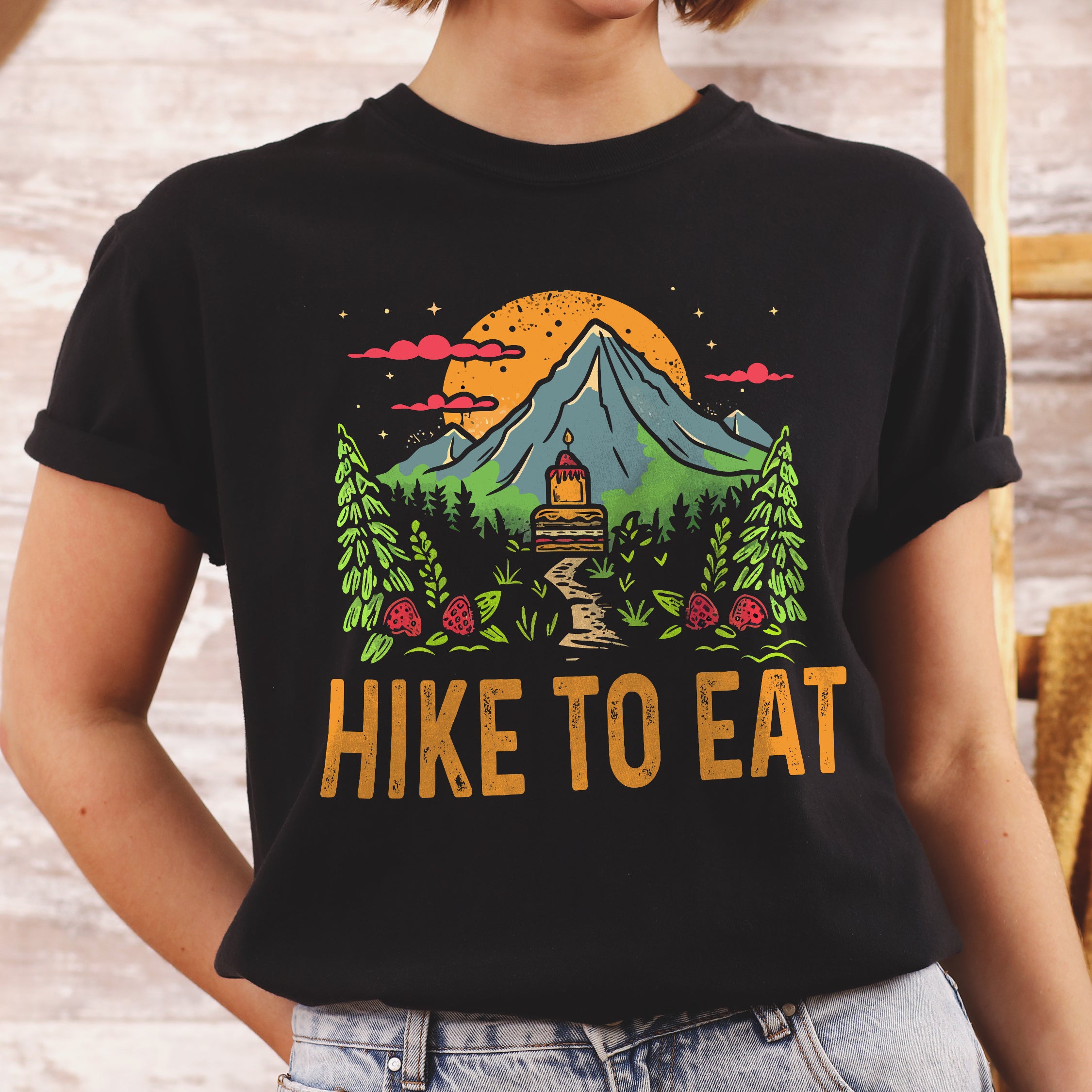 Black graphic t-shirt with 'Hike to Eat' text and 'Mountain Sweets' design featuring layered cake in mountain landscape with orange sunset, pine trees, mushrooms, and starry sky worn by a woman.