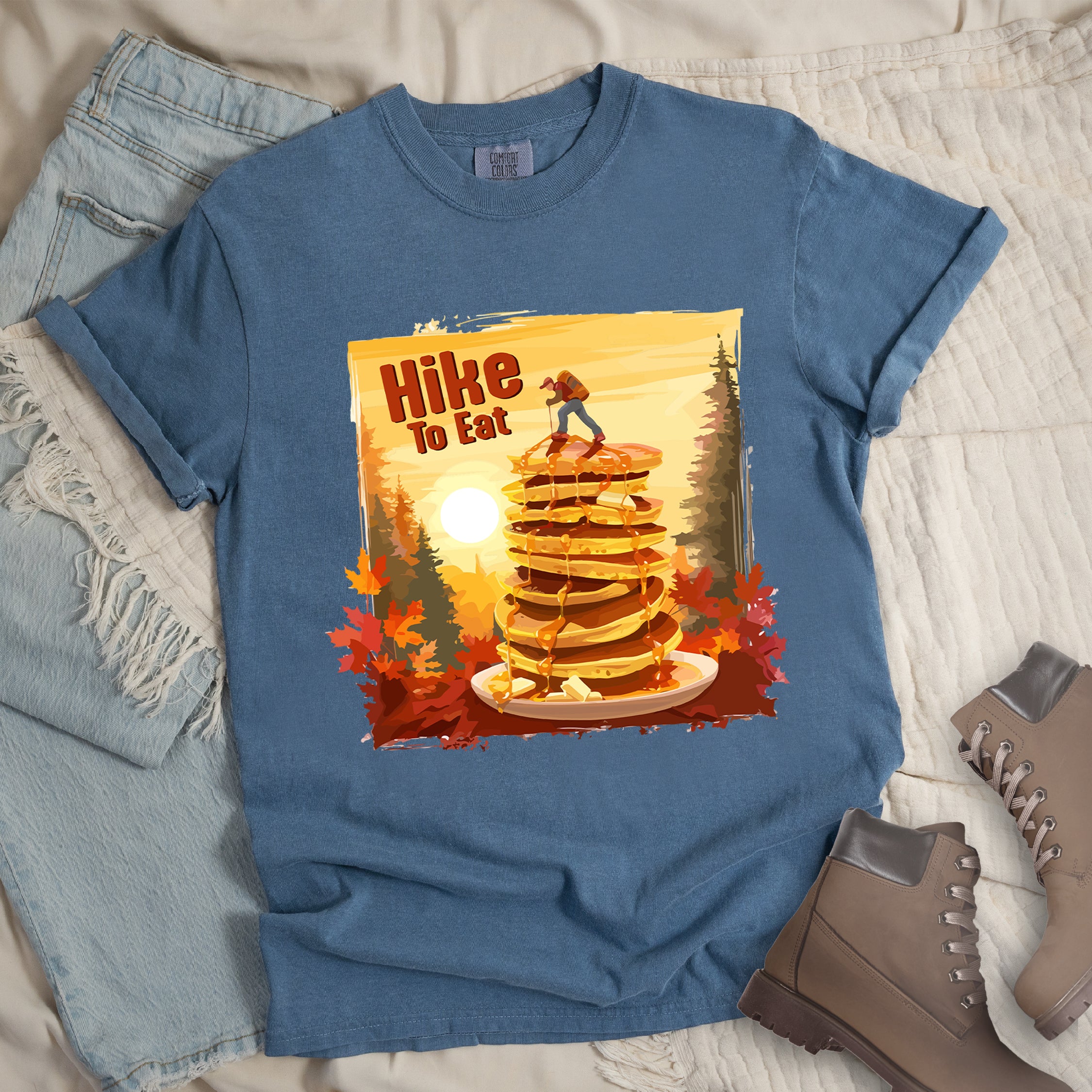 Blue Jean (Denim Blue) graphic t-shirt with 'Hike to Eat' text and 'Pancake Summit' design featuring tall stack of pancakes with hiker figure on top, set against autumn forest landscape with orange sunset, styled with light wash jeans and brown hiking boots.