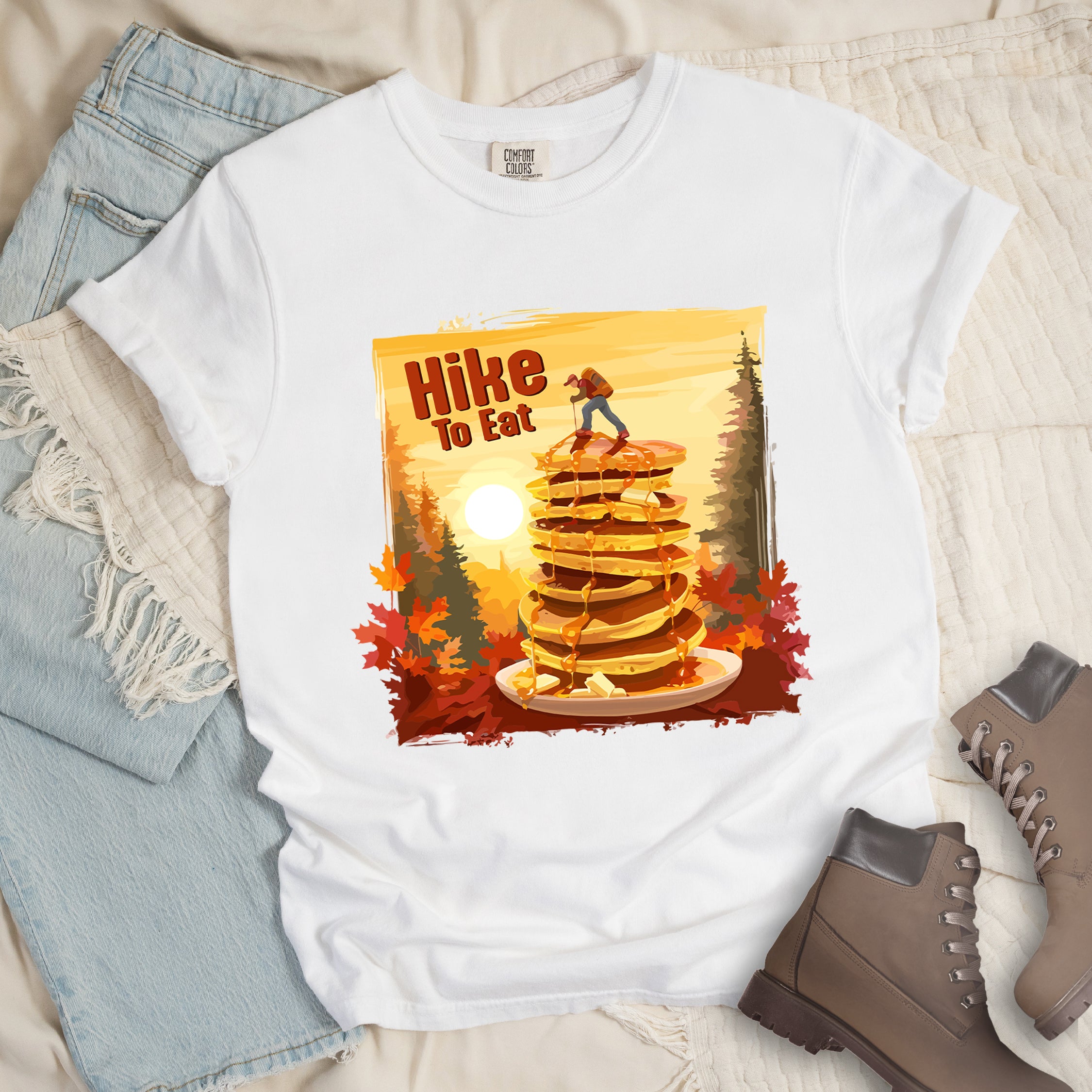 White graphic t-shirt with 'Hike to Eat' text and 'Pancake Summit' design featuring tall stack of pancakes with hiker figure on top, set against autumn forest landscape with orange sunset, styled with light wash jeans and brown hiking boots.