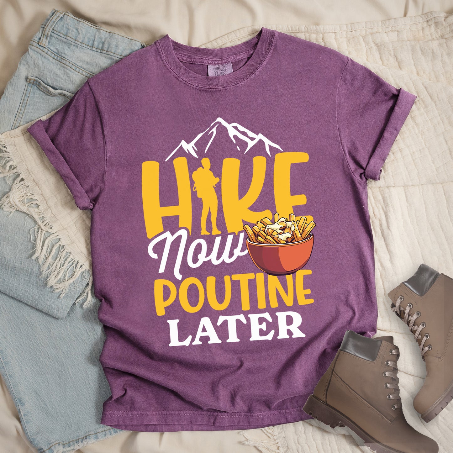 Berry (Dark Violet) t-shirt with "HIKE NOW POUTINE LATER" text design. White mountain silhouettes appear above yellow "HIKE" letters, with a hiker silhouette replacing the "I". "Now" appears in white script, "POUTINE" in yellow capital letters, and "LATER" in white. An illustration of a bowl of poutine with french fries, cheese curds and gravy appears next to "Now" and above "POUTINE". Features a classic crew neck and casual rolled sleeves, styled alongside light wash denim and brown hiking boots.