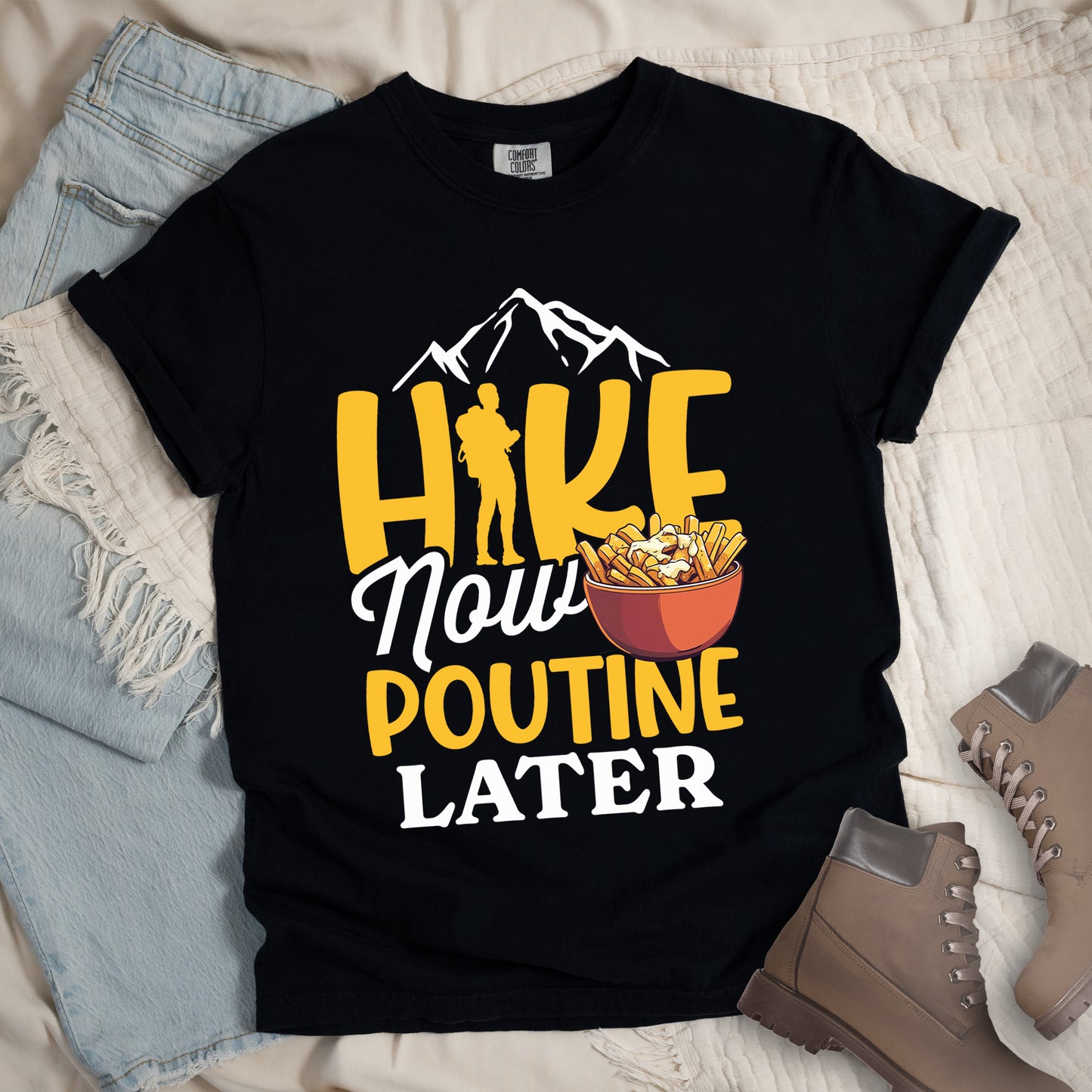 Black t-shirt with "HIKE NOW POUTINE LATER" text design. White mountain silhouettes appear above yellow "HIKE" letters, with a hiker silhouette replacing the "I". "Now" appears in white script, "POUTINE" in yellow capital letters, and "LATER" in white. An illustration of a bowl of poutine with french fries, cheese curds and gravy appears next to "Now" and above "POUTINE". Features a classic crew neck and casual rolled sleeves, styled alongside light wash denim and brown hiking boots.