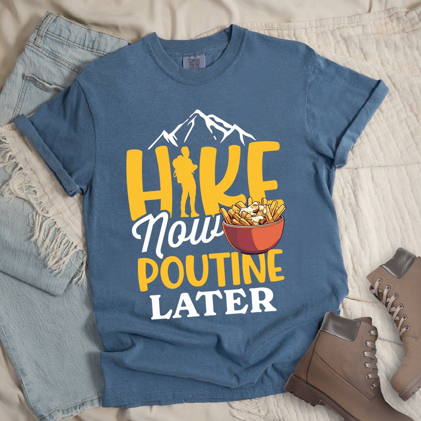 Blue Jean (Denim Blue) t-shirt with "HIKE NOW POUTINE LATER" text design. White mountain silhouettes appear above yellow "HIKE" letters, with a hiker silhouette replacing the "I". "Now" appears in white script, "POUTINE" in yellow capital letters, and "LATER" in white. An illustration of a bowl of poutine with french fries, cheese curds and gravy appears next to "Now" and above "POUTINE". Features a classic crew neck and casual rolled sleeves, styled alongside light wash denim and brown hiking boots.
