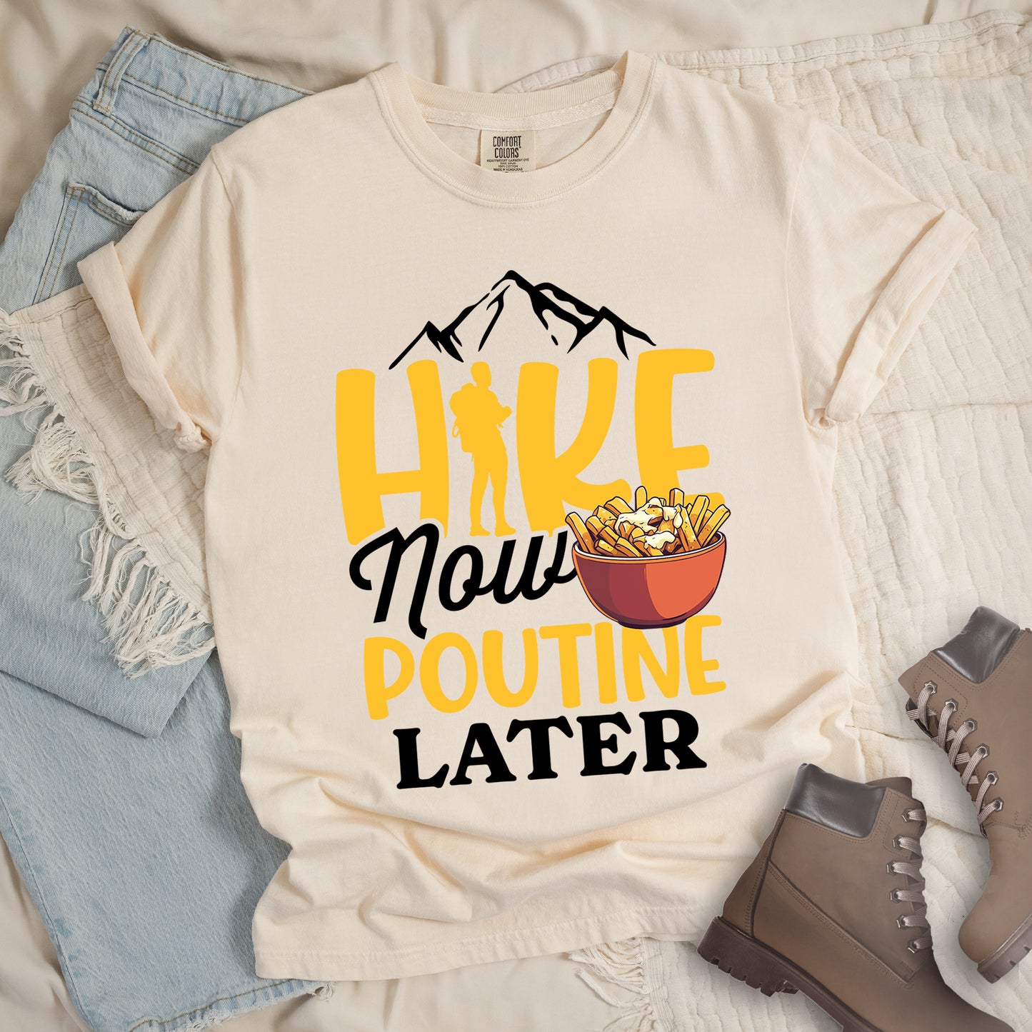 Ivory (Cream White) t-shirt with "HIKE NOW POUTINE LATER" text design. Black mountain silhouettes appear above yellow "HIKE" letters, with a hiker silhouette replacing the "I". "Now" appears in black script, "POUTINE" in yellow capital letters, and "LATER" in black. An illustration of a bowl of poutine with french fries, cheese curds and gravy appears next to "Now" and above "POUTINE". Features a classic crew neck and casual rolled sleeves, styled alongside light wash denim and brown hiking boots.