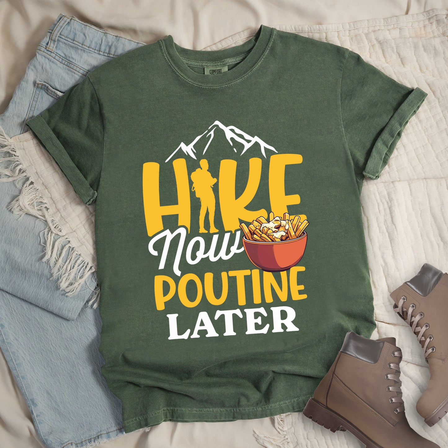 Moss (Dark Green) t-shirt with "HIKE NOW POUTINE LATER" text design. White mountain silhouettes appear above yellow "HIKE" letters, with a hiker silhouette replacing the "I". "Now" appears in white script, "POUTINE" in yellow capital letters, and "LATER" in white. An illustration of a bowl of poutine with french fries, cheese curds and gravy appears next to "Now" and above "POUTINE". Features a classic crew neck and casual rolled sleeves, styled alongside light wash denim and brown hiking boots.