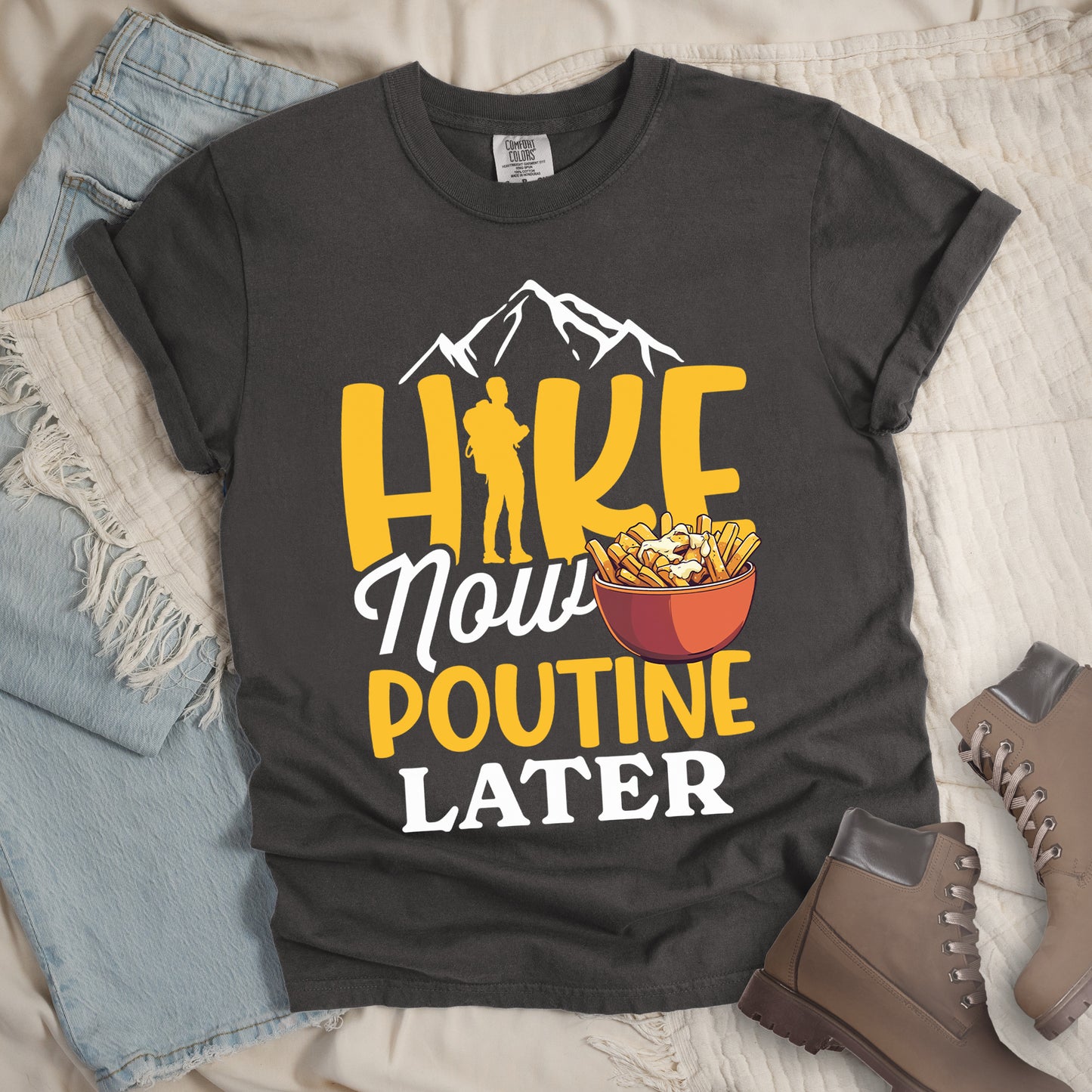 Pepper (Dark Gray) t-shirt with "HIKE NOW POUTINE LATER" text design. White mountain silhouettes appear above yellow "HIKE" letters, with a hiker silhouette replacing the "I". "Now" appears in white script, "POUTINE" in yellow capital letters, and "LATER" in white. An illustration of a bowl of poutine with french fries, cheese curds and gravy appears next to "Now" and above "POUTINE". Features a classic crew neck and casual rolled sleeves, styled alongside light wash denim and brown hiking boots.