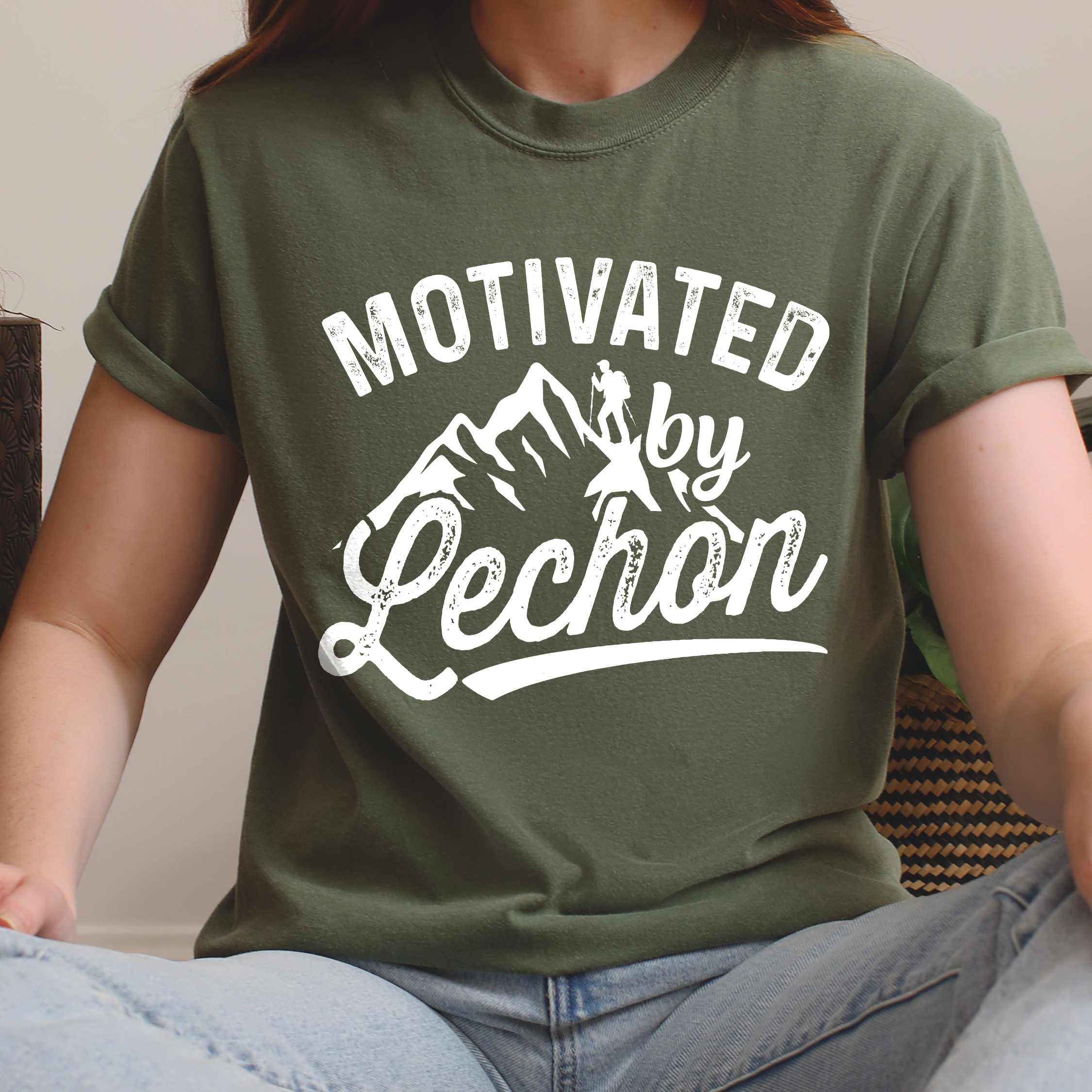 Woman wearing moss colored graphic t-shirt with 'Motivated by Lechon' text in white lettering featuring mountain silhouettes and hiker illustration
