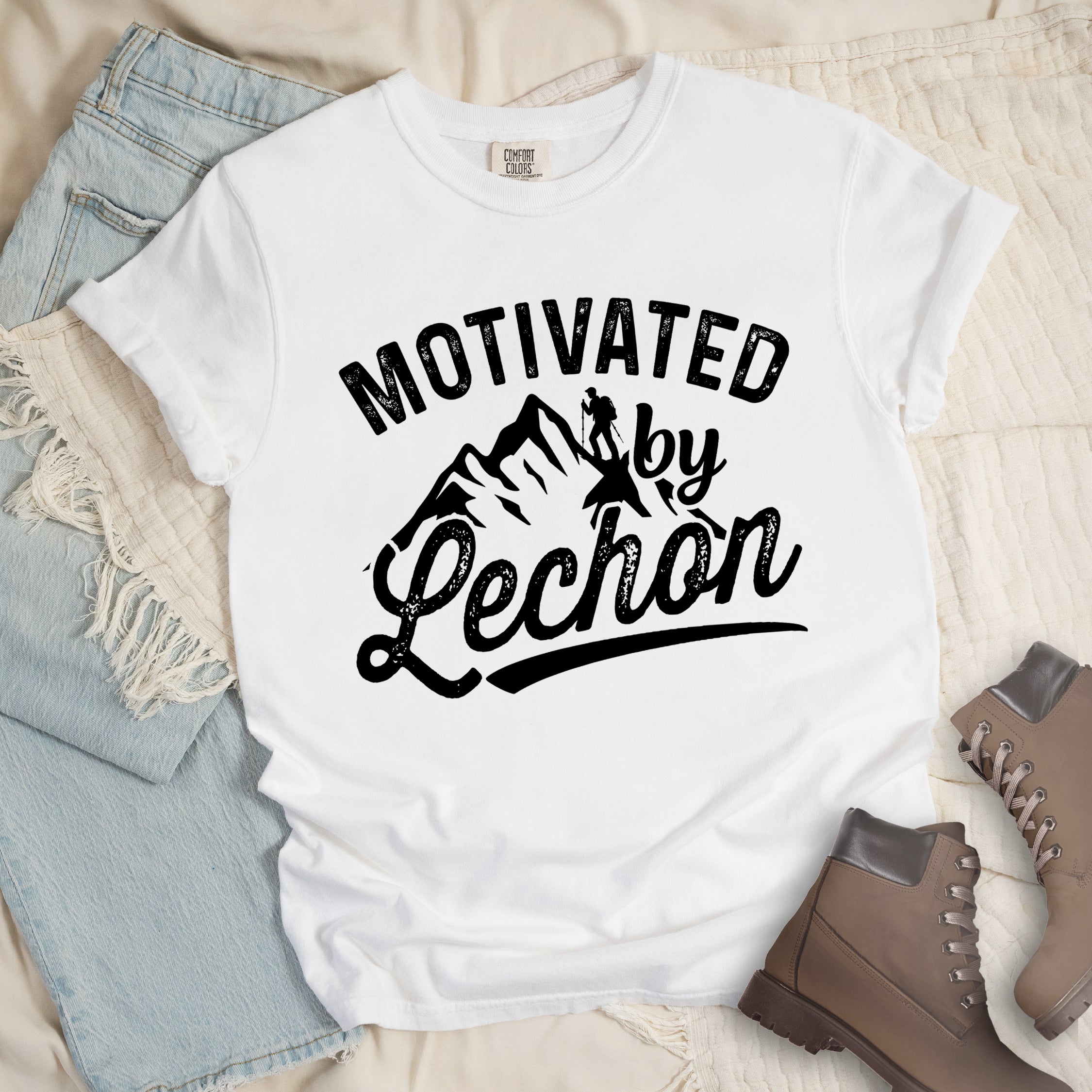 White graphic t-shirt with 'Motivated by Lechon' text in black lettering featuring mountain silhouettes and hiker illustration