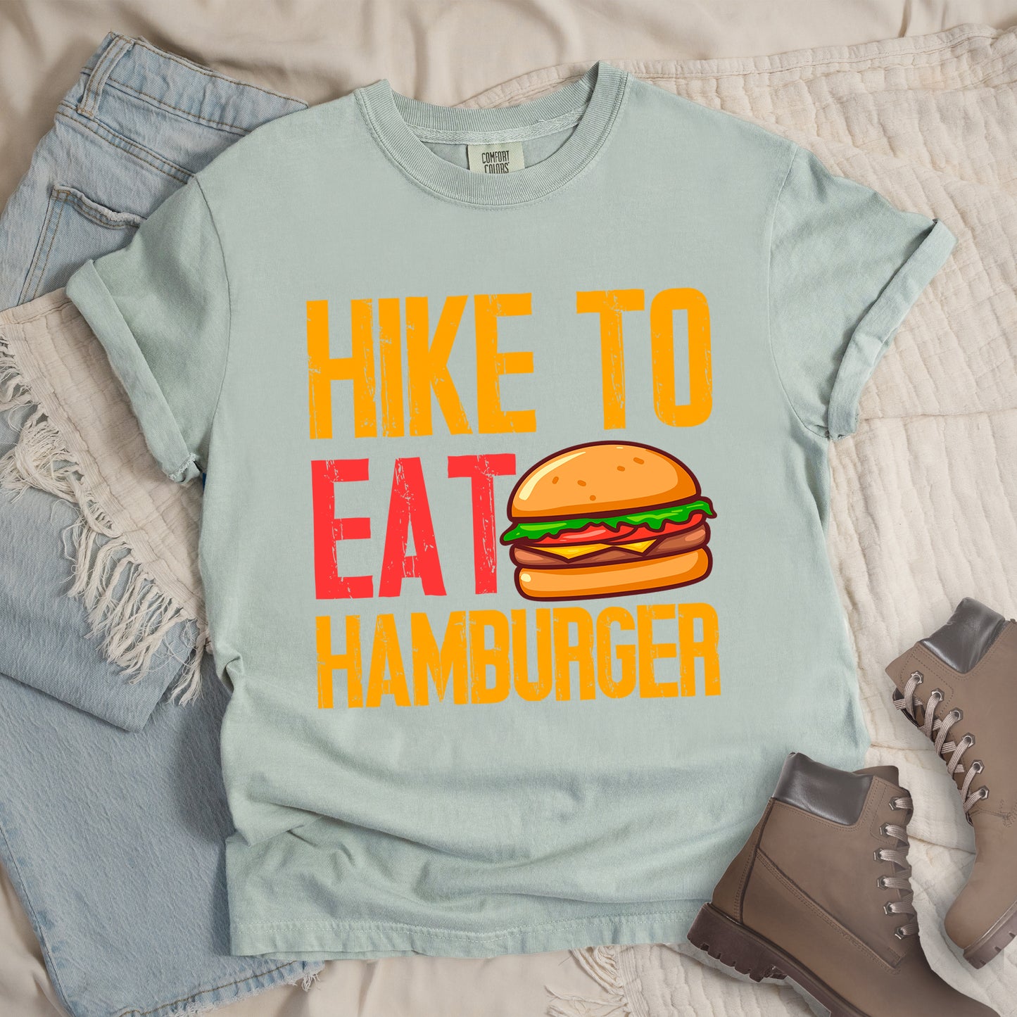 Bay (Light Gray) t-shirt with distressed text reading "HIKE TO EAT HAMBURGER" arranged in three lines. "HIKE TO" appears in yellow at the top, "EAT" in red in the middle with a colorful hamburger illustration next to it, and "HAMBURGER" in yellow at the bottom. The hamburger graphic shows a bun, meat patty, cheese, lettuce, and tomato. Features a classic crew neck and casual rolled sleeves, styled alongside light wash denim and brown hiking boots.