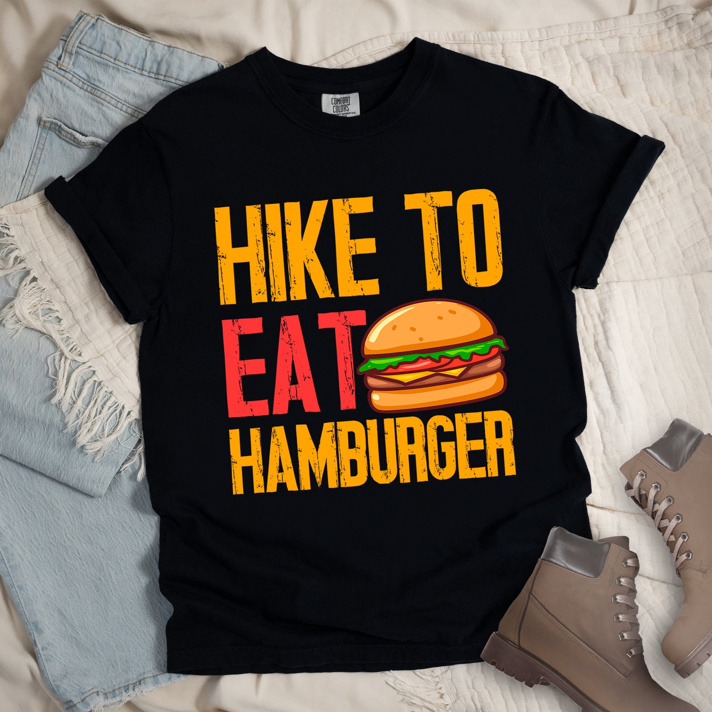 Black t-shirt with distressed text reading "HIKE TO EAT HAMBURGER" arranged in three lines. "HIKE TO" appears in yellow at the top, "EAT" in red in the middle with a colorful hamburger illustration next to it, and "HAMBURGER" in yellow at the bottom. The hamburger graphic shows a bun, meat patty, cheese, lettuce, and tomato. Features a classic crew neck and casual rolled sleeves, styled alongside light wash denim and brown hiking boots.