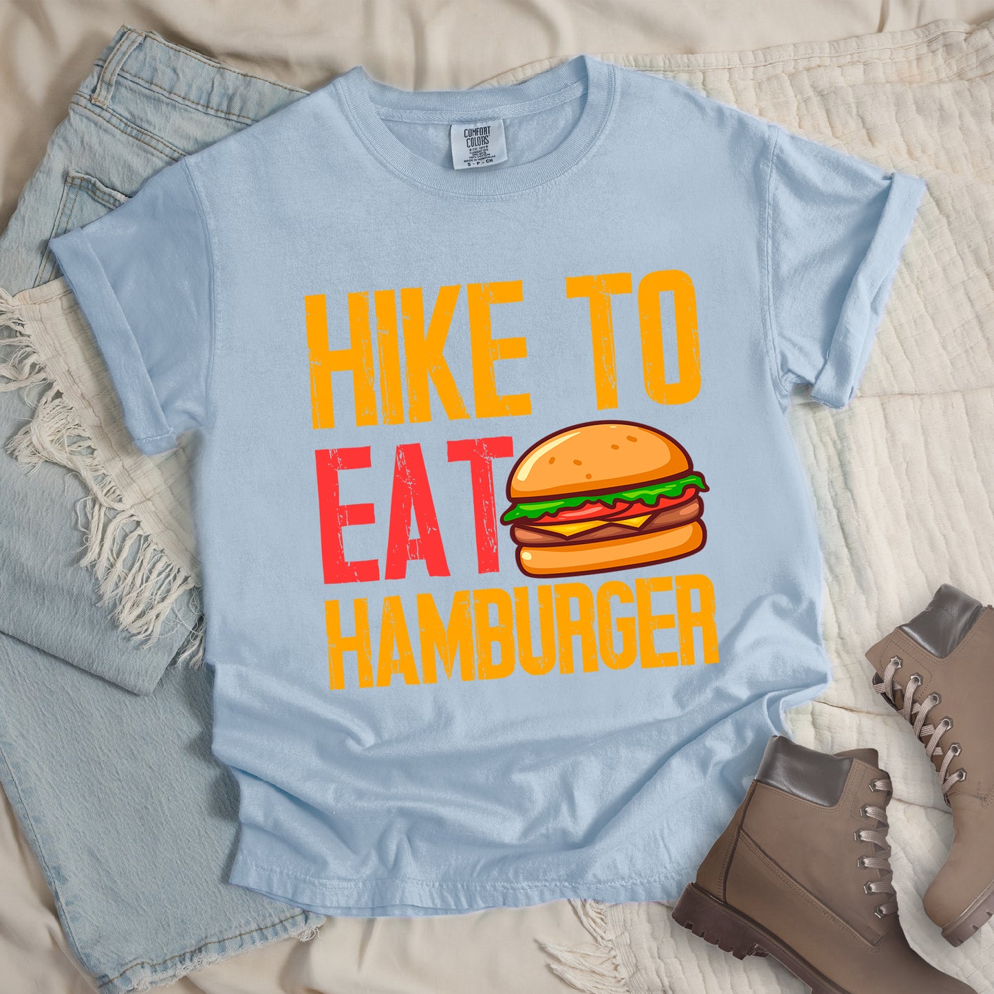 Chambray (Light Blue) t-shirt with distressed text reading "HIKE TO EAT HAMBURGER" arranged in three lines. "HIKE TO" appears in yellow at the top, "EAT" in red in the middle with a colorful hamburger illustration next to it, and "HAMBURGER" in yellow at the bottom. The hamburger graphic shows a bun, meat patty, cheese, lettuce, and tomato. Features a classic crew neck and casual rolled sleeves, styled alongside light wash denim and brown hiking boots.