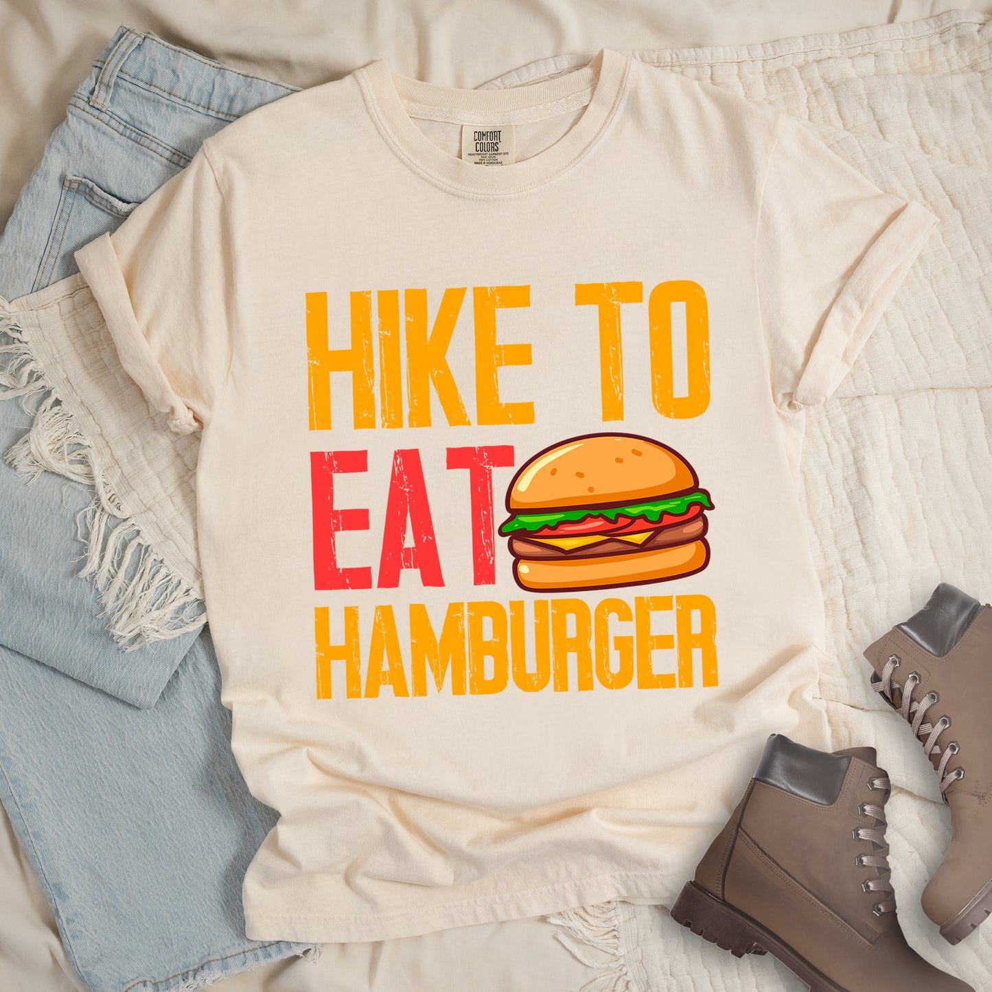 Ivory (Cream White) t-shirt with distressed text reading "HIKE TO EAT HAMBURGER" arranged in three lines. "HIKE TO" appears in yellow at the top, "EAT" in red in the middle with a colorful hamburger illustration next to it, and "HAMBURGER" in yellow at the bottom. The hamburger graphic shows a bun, meat patty, cheese, lettuce, and tomato. Features a classic crew neck and casual rolled sleeves, styled alongside light wash denim and brown hiking boots.