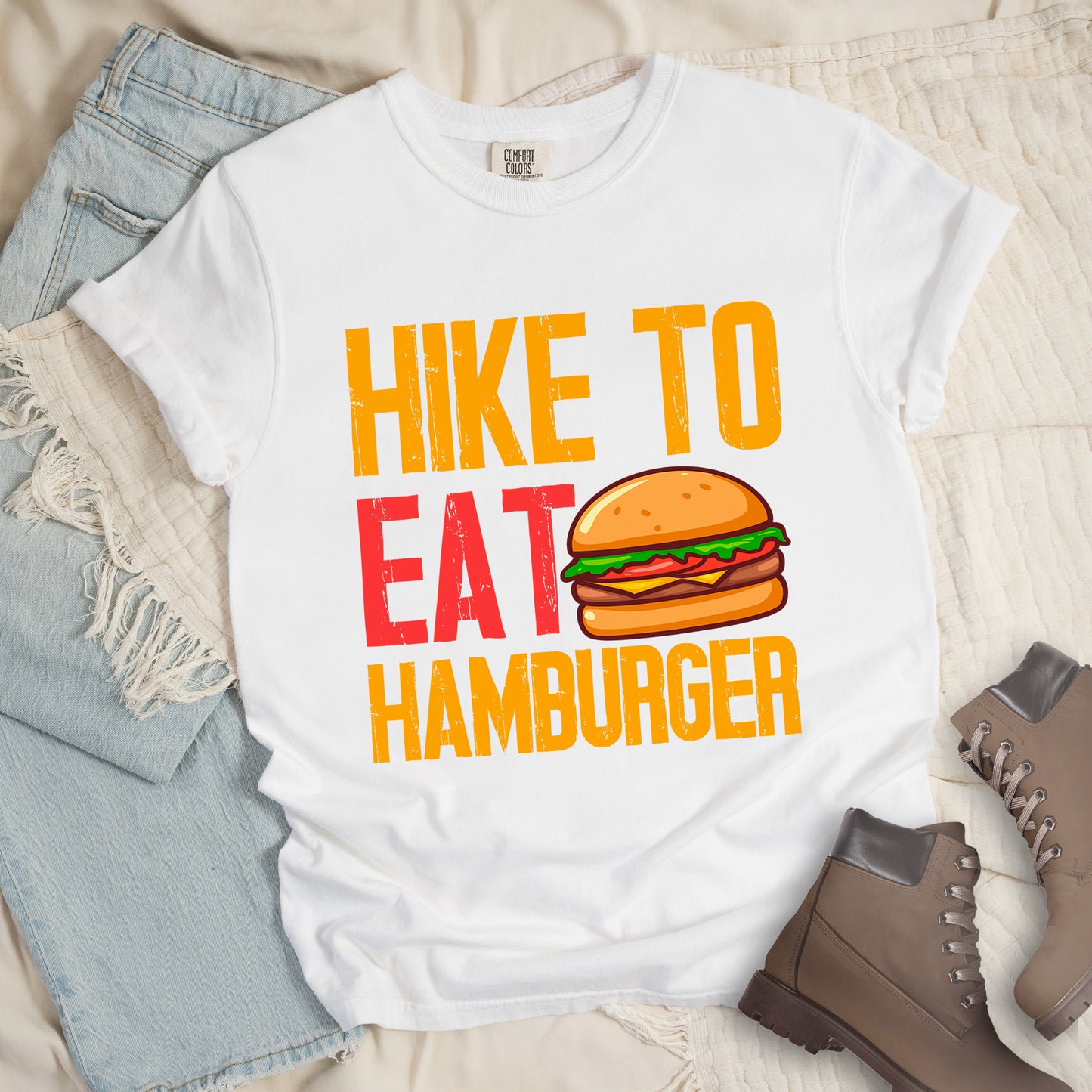 White t-shirt with distressed text reading "HIKE TO EAT HAMBURGER" arranged in three lines. "HIKE TO" appears in yellow at the top, "EAT" in red in the middle with a colorful hamburger illustration next to it, and "HAMBURGER" in yellow at the bottom. The hamburger graphic shows a bun, meat patty, cheese, lettuce, and tomato. Features a classic crew neck and casual rolled sleeves, styled alongside light wash denim and brown hiking boots.