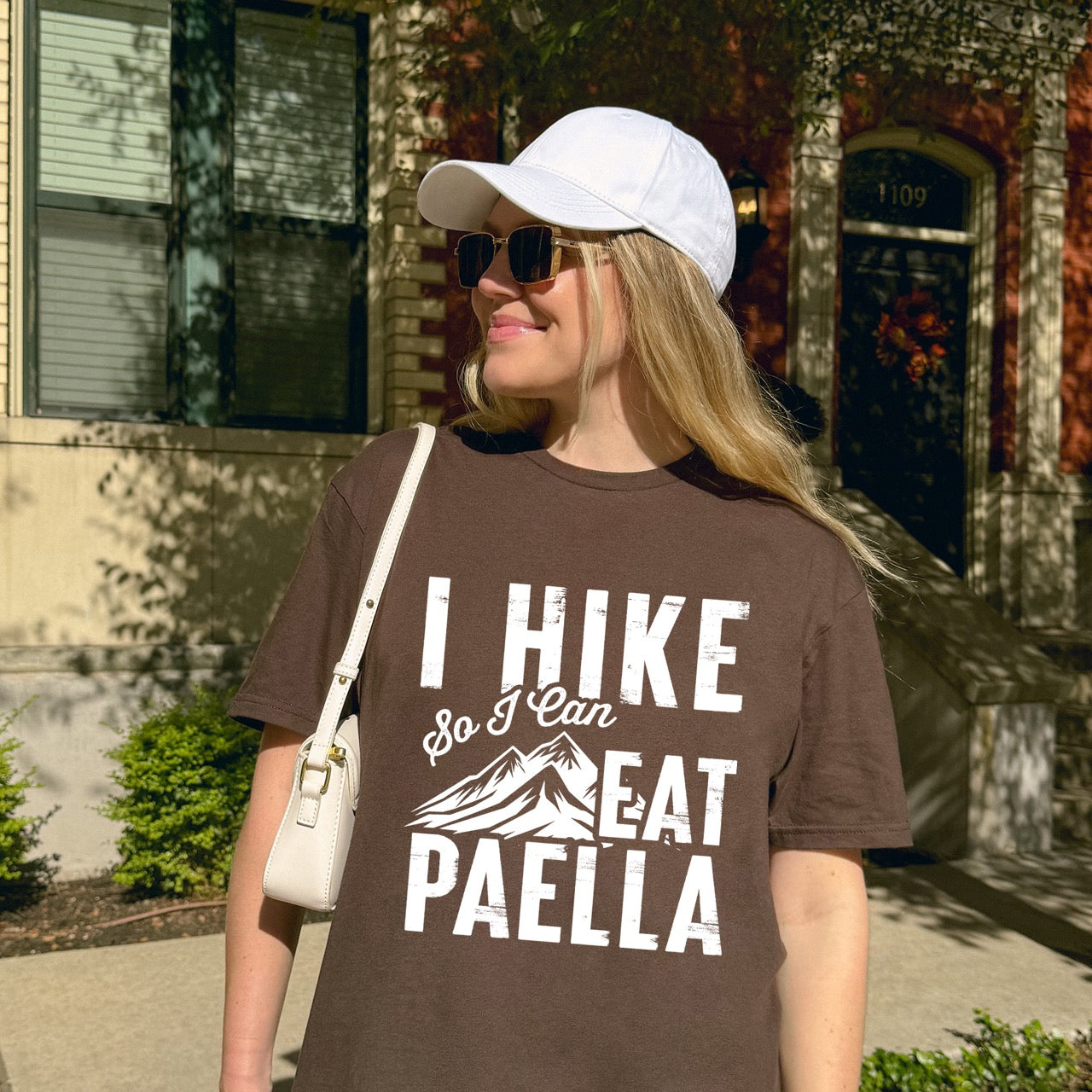 woman wearing chocolate brown Hike Now Paella Later statement tee with bold distressed typography and hiker silhouette for Spanish food-loving outdoor enthusiasts and hikers