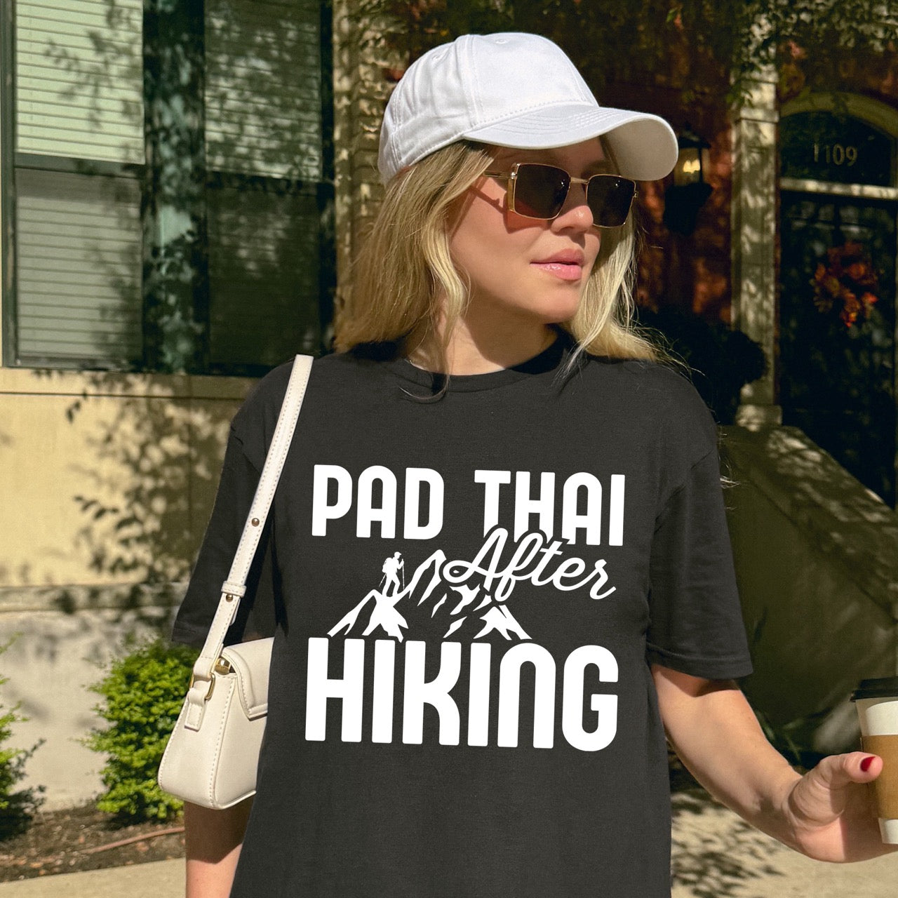 woman with glasses wearing black Pad Thai after Hiking statement tee with bold typography, elegant script lettering, mountain peaks, and hiker silhouette for Thai food-loving outdoor enthusiasts, foodies and hikers.