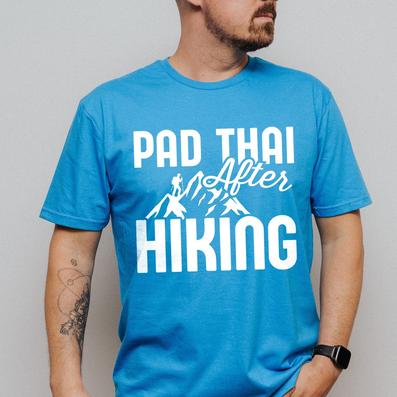 man wearing saphire (blue) Pad Thai after Hiking statement tee with bold typography, elegant script lettering, mountain peaks, and hiker silhouette for Thai food-loving outdoor enthusiasts, foodies and hikers.
