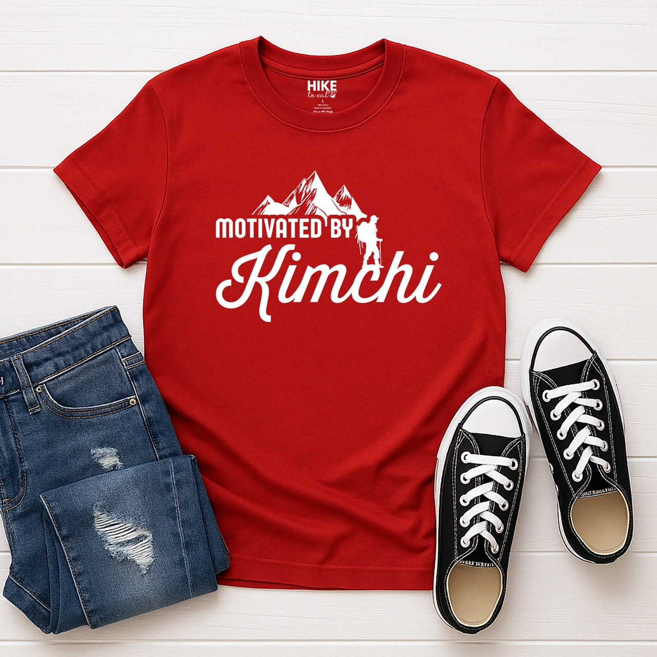 red Motivated by Kimchi statement tee with bold typography, elegant script lettering, mountain peaks, and hiker silhouette for Korean food-loving outdoor enthusiasts, foodies and hikers laid flat with shoes and jeans