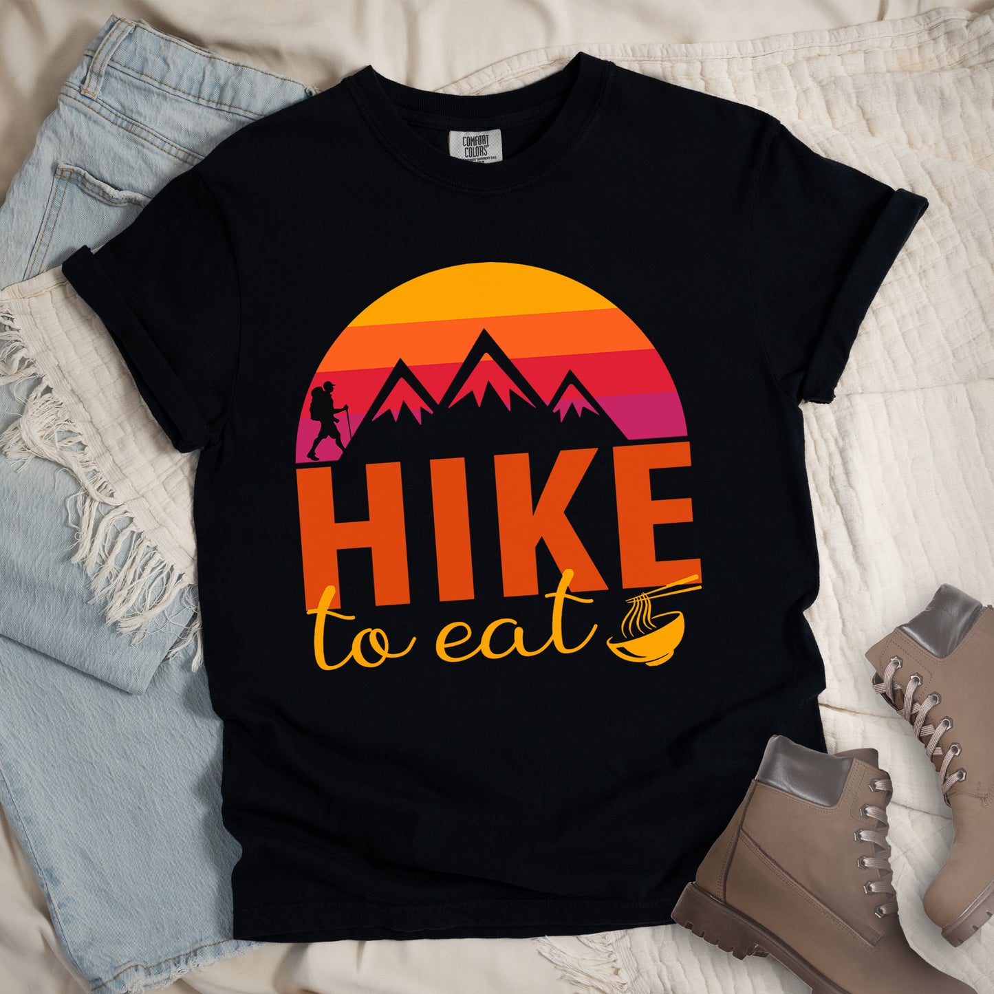 Black t-shirt featuring a retro sunset design with gradient colors from yellow to orange to red to purple. Mountain silhouettes and a small hiker figure appear against the sunset. Below this is "HIKE" in large orange letters and "to eat" in yellow script, followed by a small illustration of a noodle bowl with chopsticks. The design has a vintage outdoor aesthetic. The shirt features a classic crew neck and casual rolled sleeves, styled alongside light wash denim and brown hiking boots.