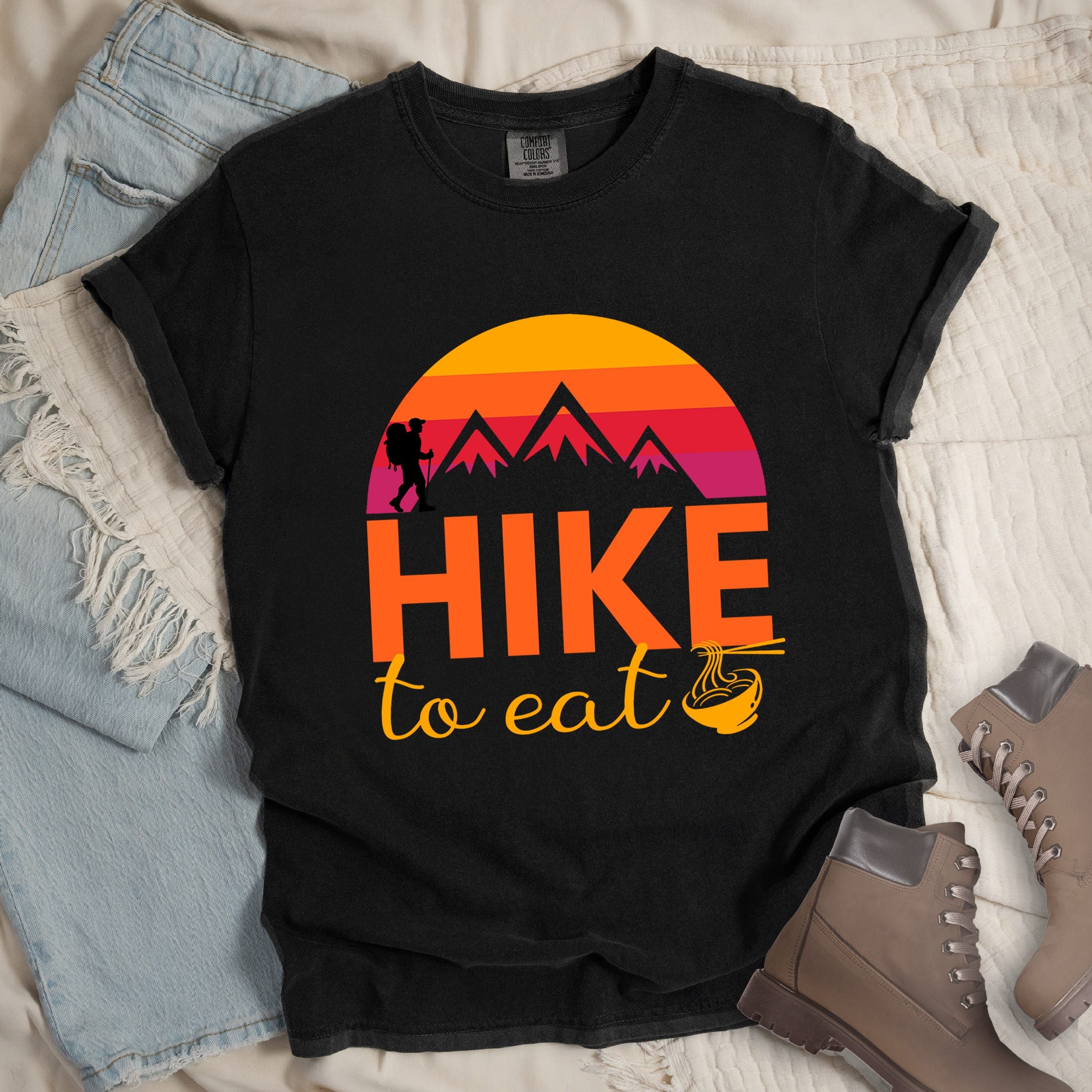 Black shirt with the text “Hike to Eat” and a circular mountain and sunset graphic, with a small bowl of noodles integrated into the design.