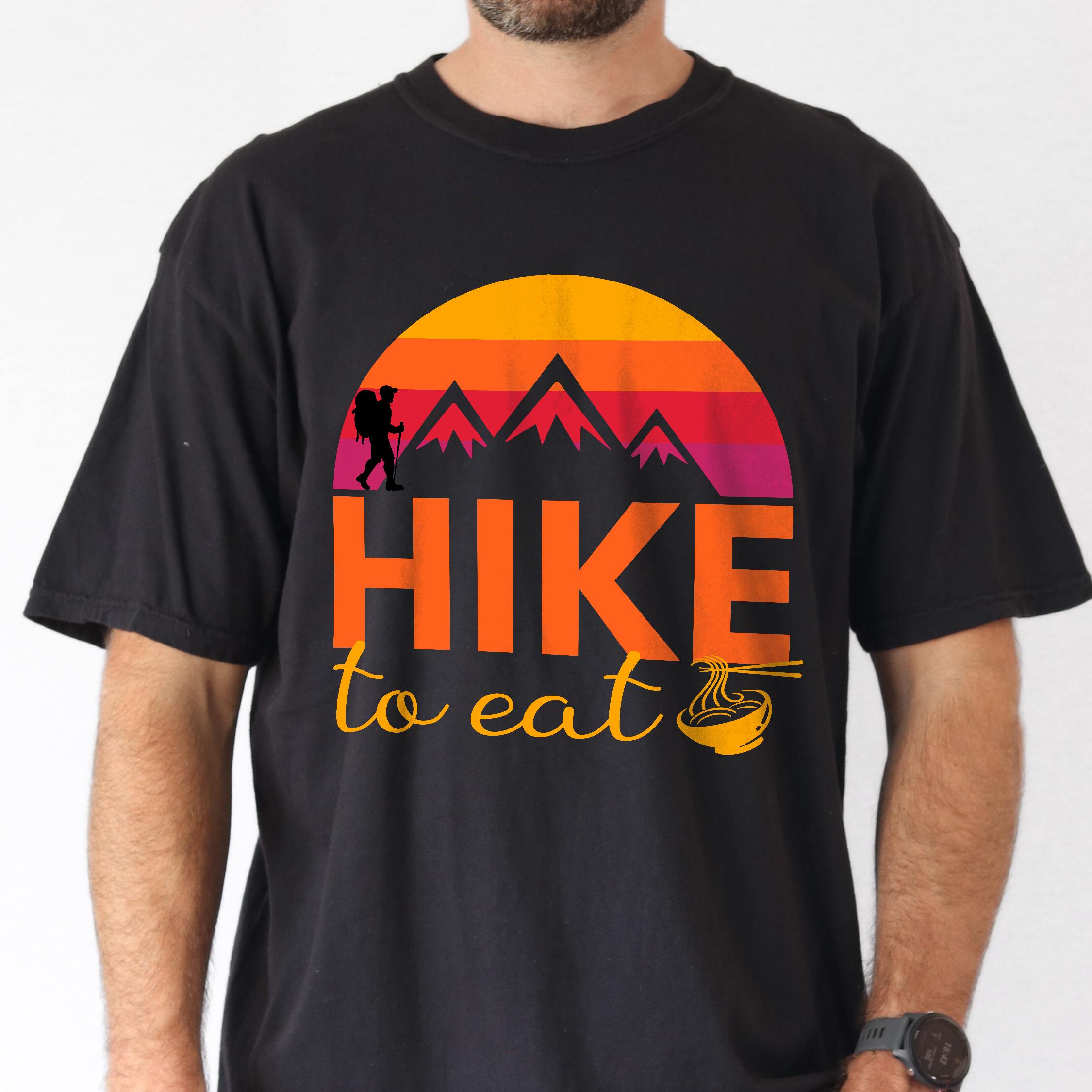 Man wearing black "Hike to Eat" colored vintage tee with bold typography, mountains, sunrise, and backpacker to savor each step along the trail.