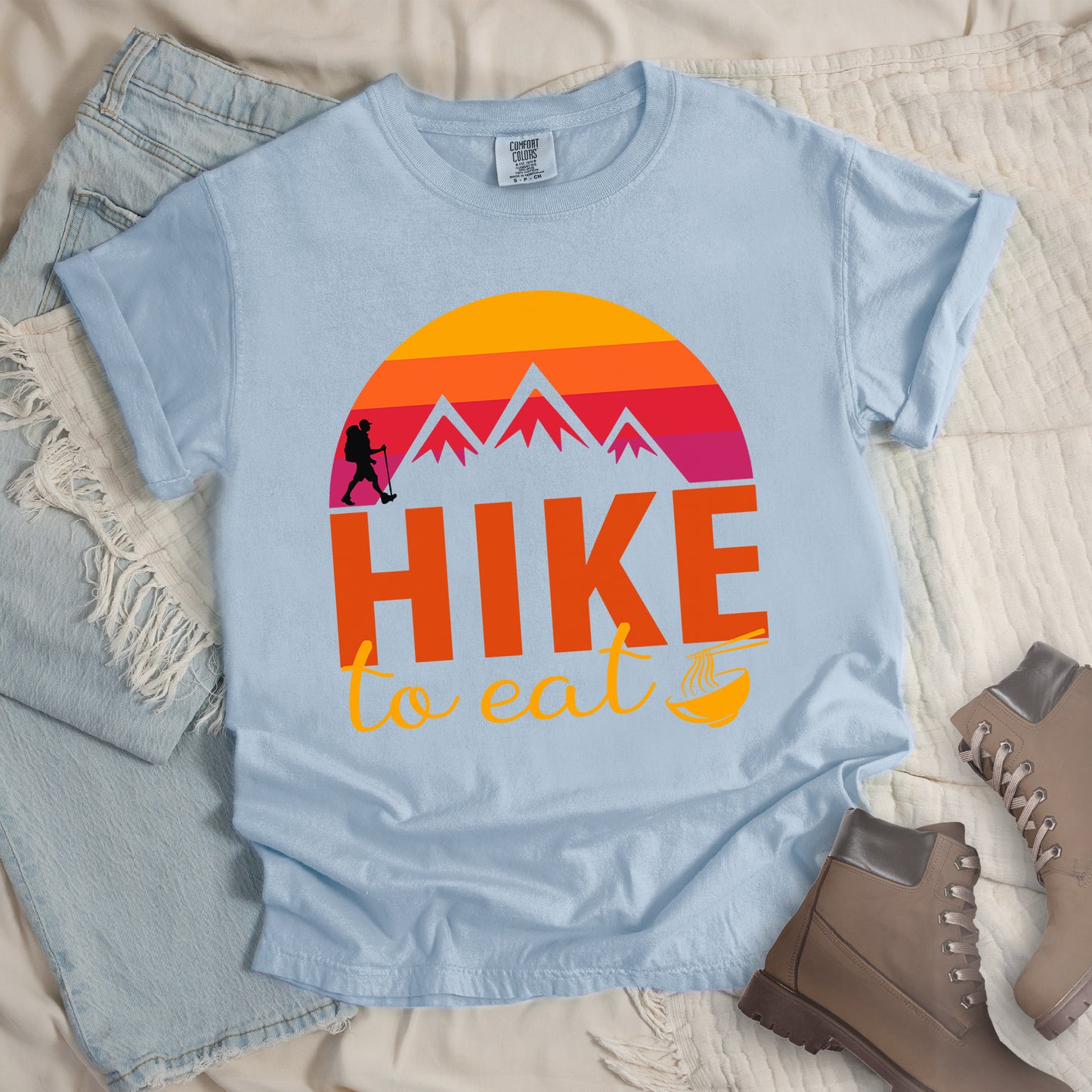 Chambray (Light Blue) t-shirt featuring a retro sunset design with gradient colors from yellow to orange to red to purple. Mountain silhouettes and a small hiker figure appear against the sunset. Below this is "HIKE" in large orange letters and "to eat" in yellow script, followed by a small illustration of a noodle bowl with chopsticks. The design has a vintage outdoor aesthetic. The shirt features a classic crew neck and casual rolled sleeves, styled alongside light wash denim and brown hiking boots.