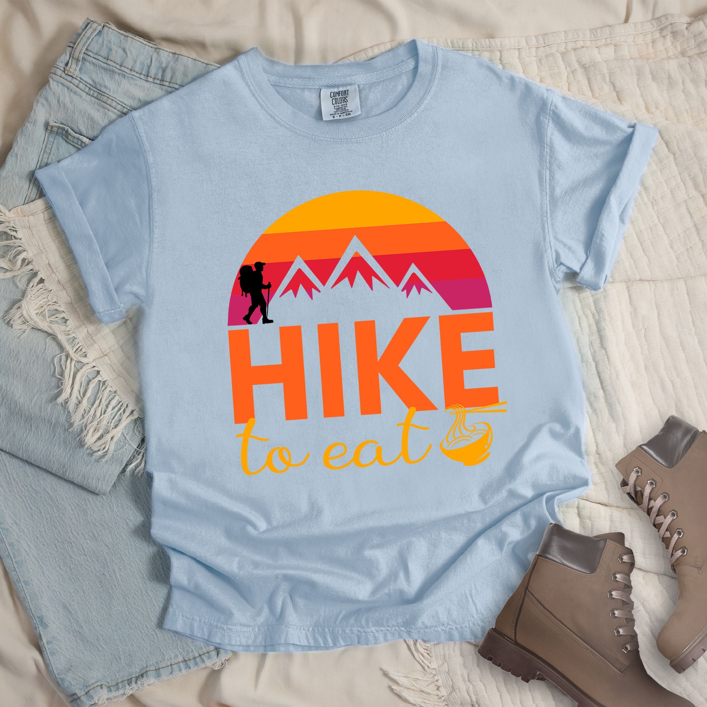Light blue shirt with the text “Hike to Eat” and a circular mountain and sunset graphic, with a small bowl of noodles integrated into the design.