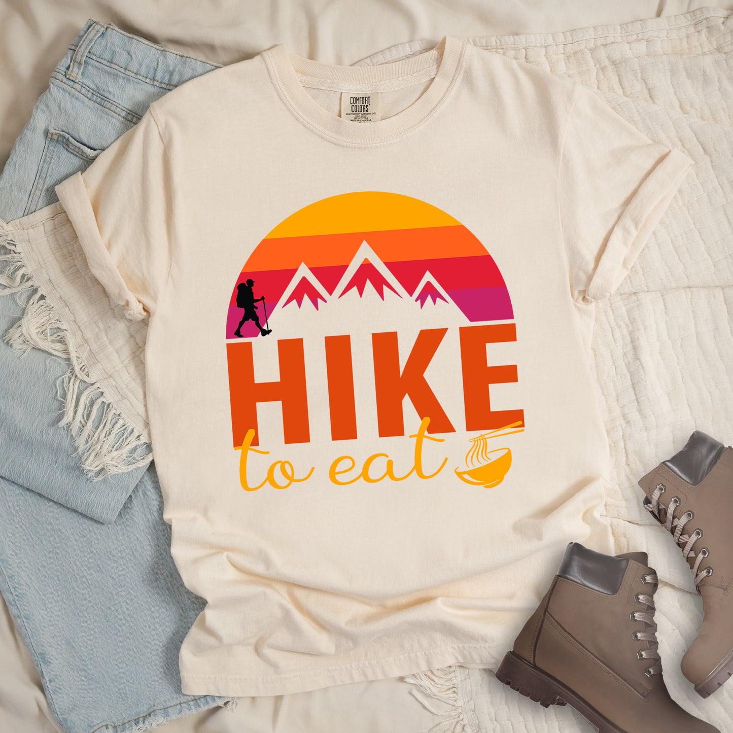 Ivory (Cream White) t-shirt featuring a retro sunset design with gradient colors from yellow to orange to red to purple. Mountain silhouettes and a small hiker figure appear against the sunset. Below this is "HIKE" in large orange letters and "to eat" in yellow script, followed by a small illustration of a noodle bowl with chopsticks. The design has a vintage outdoor aesthetic. The shirt features a classic crew neck and casual rolled sleeves, styled alongside light wash denim and brown hiking boots.