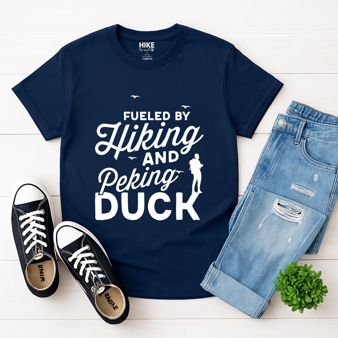 Navy Fueled by Hiking and Peking Duck statement tee with bold and script typography, flying birds, and hiker silhouette for Chinese food-loving outdoor enthusiasts, hikers and foodies laid flat with jeans and shoes