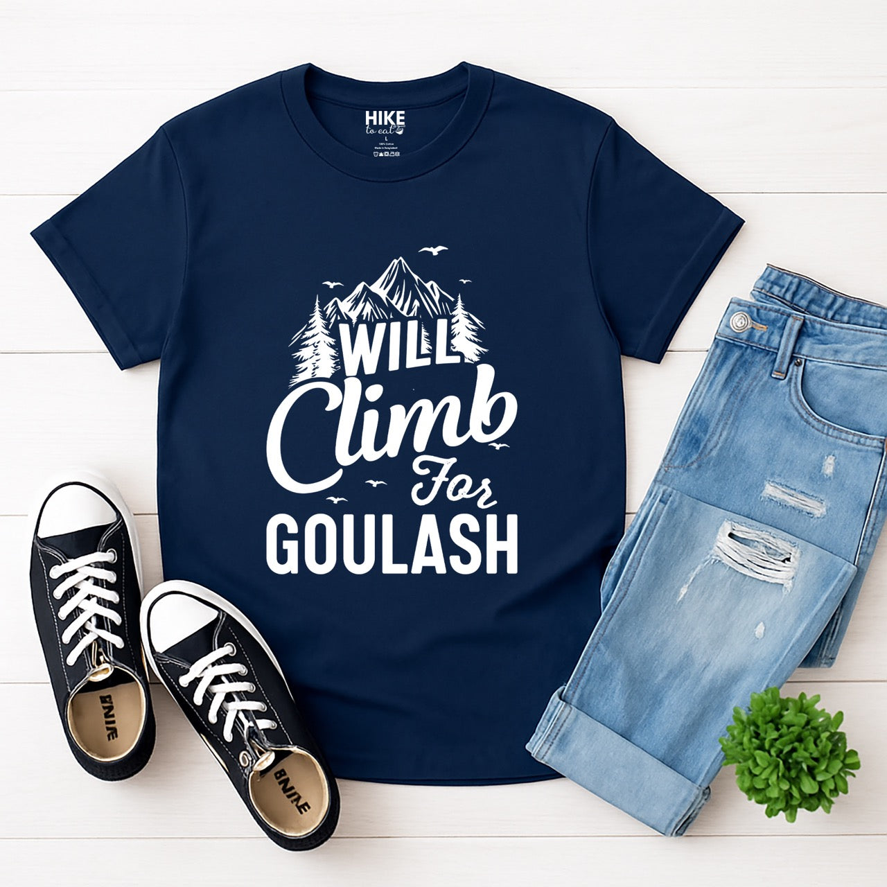 Navy Will Climb for Goulash statement tee with bold and script typography, mountain peaks, pine trees, and flying birds for Hungarian food-loving climbers, hikers and foodies laid flat with jeans and shoes