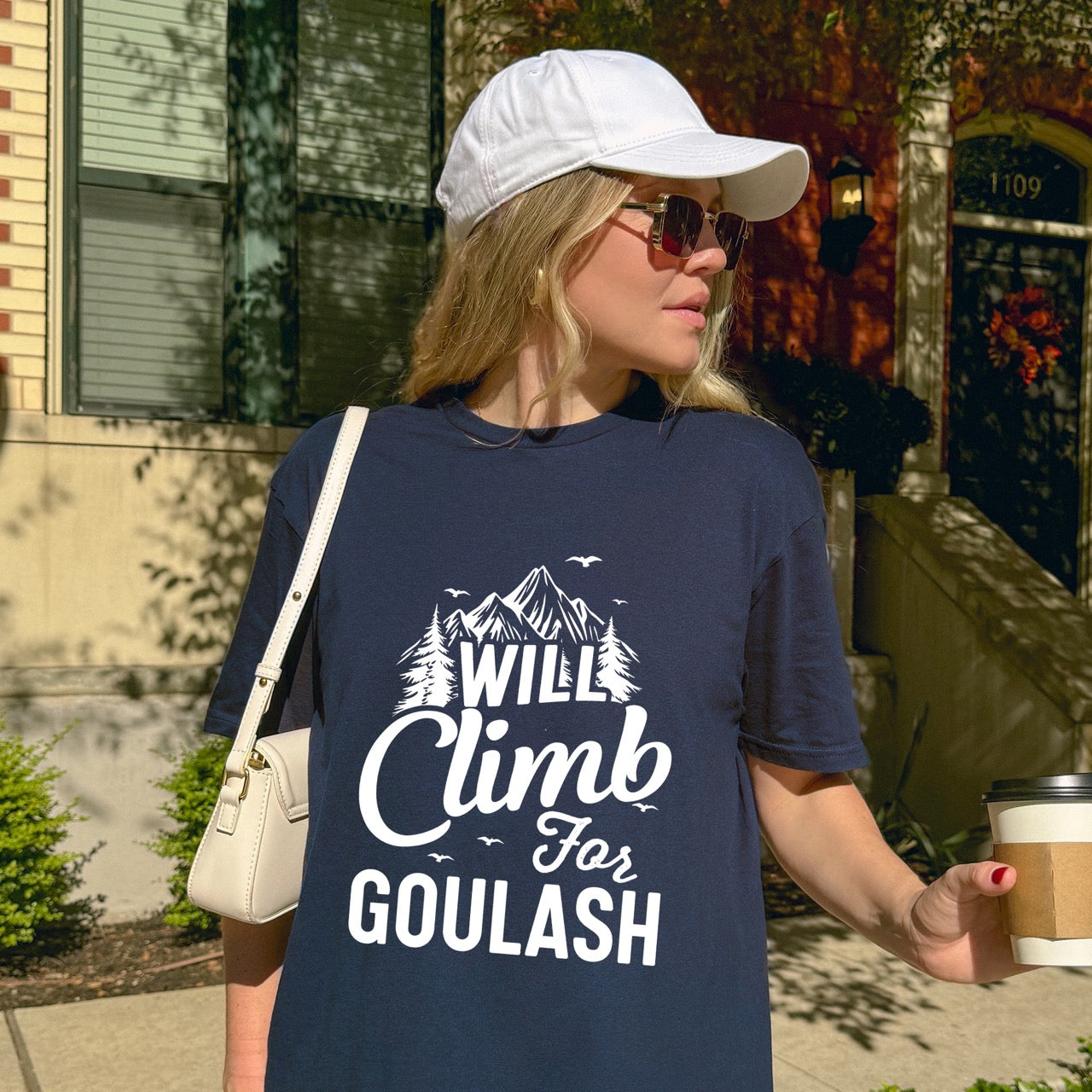 woman wearing Navy Will Climb for Goulash statement tee with bold and script typography, mountain peaks, pine trees, and flying birds for Hungarian food-loving climbers, hikers and foodies