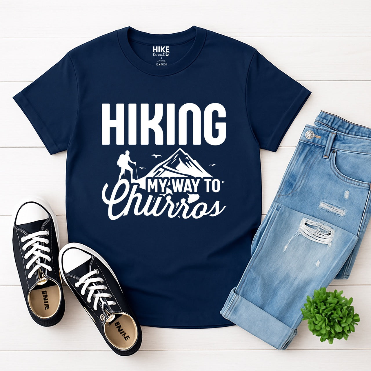 Navy Hiking my way to Churros statement tee with bold typography, script lettering, mountain peaks, hiker silhouette, and flying birds for Spanish dessert-loving outdoor enthusiasts, hikers and foodies laid flat with shoes and jeans