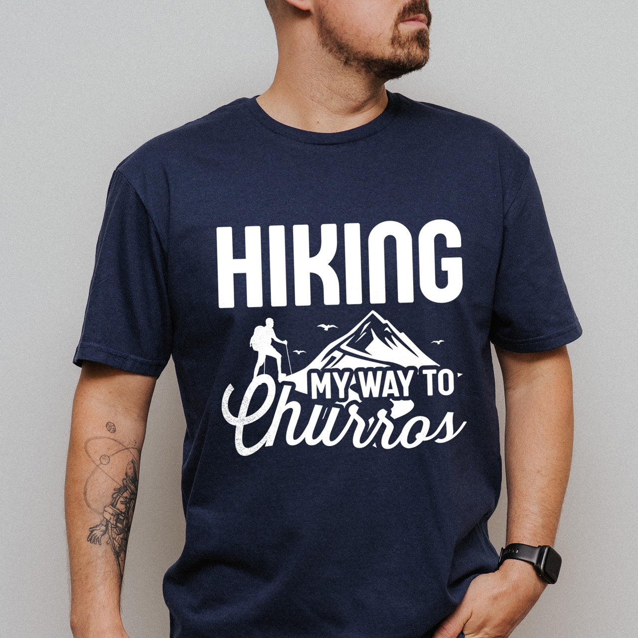 Man wearing Navy Hiking my way to Churros statement tee with bold typography, script lettering, mountain peaks, hiker silhouette, and flying birds for Spanish dessert-loving outdoor enthusiasts, hikers and foodies
