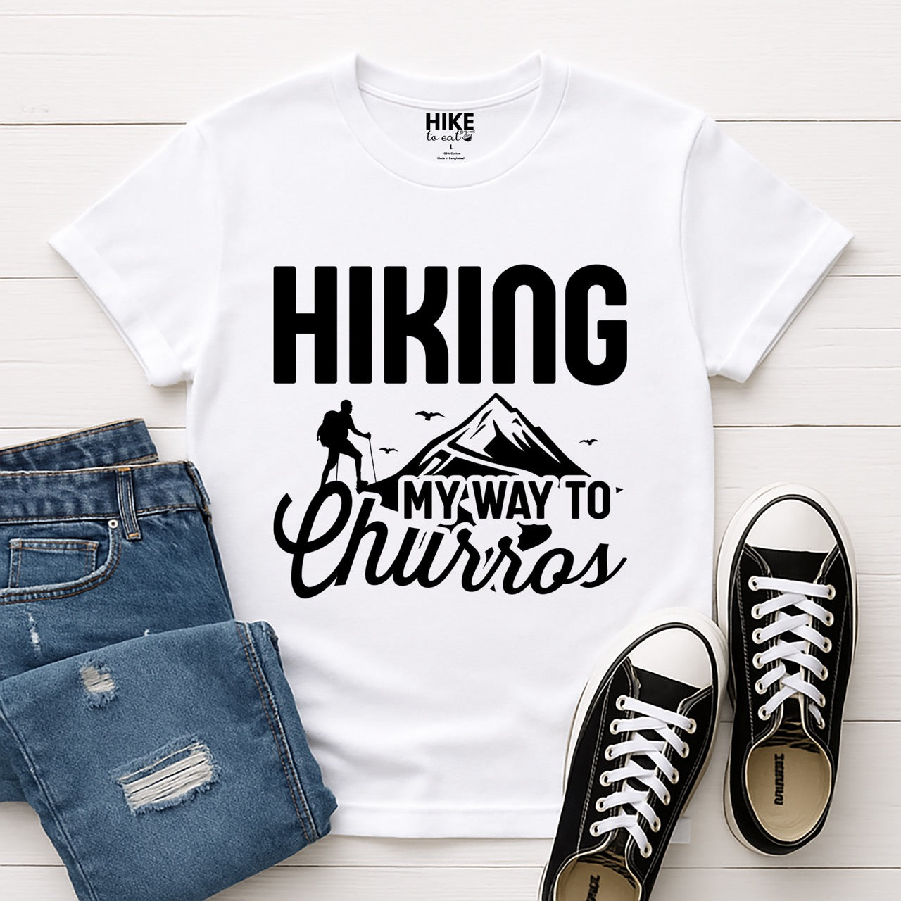 white Hiking my way to Churros statement tee with bold typography, script lettering, mountain peaks, hiker silhouette, and flying birds for Spanish dessert-loving outdoor enthusiasts, hikers and foodies laid flat with shoes and jeans