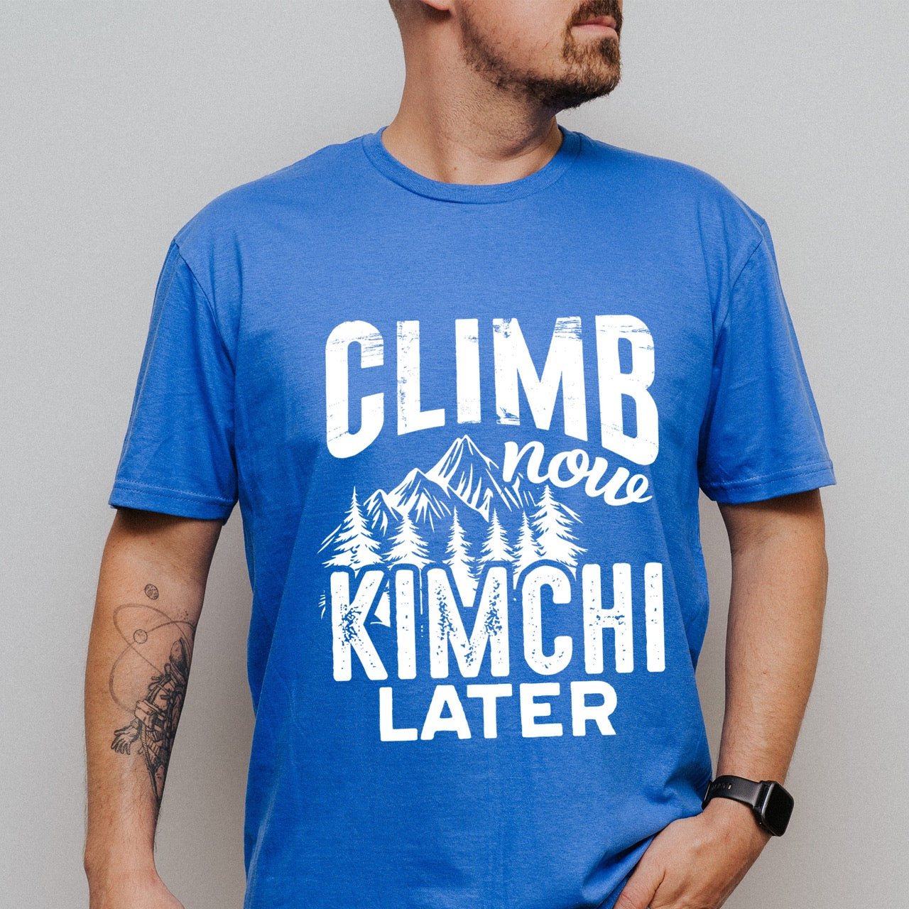 Man wearing Royal blue Climb now Kimchi later statement tee with distressed bold typography, script lettering, mountain peaks, and pine trees for Korean food-loving climbers, hikers and foodies.