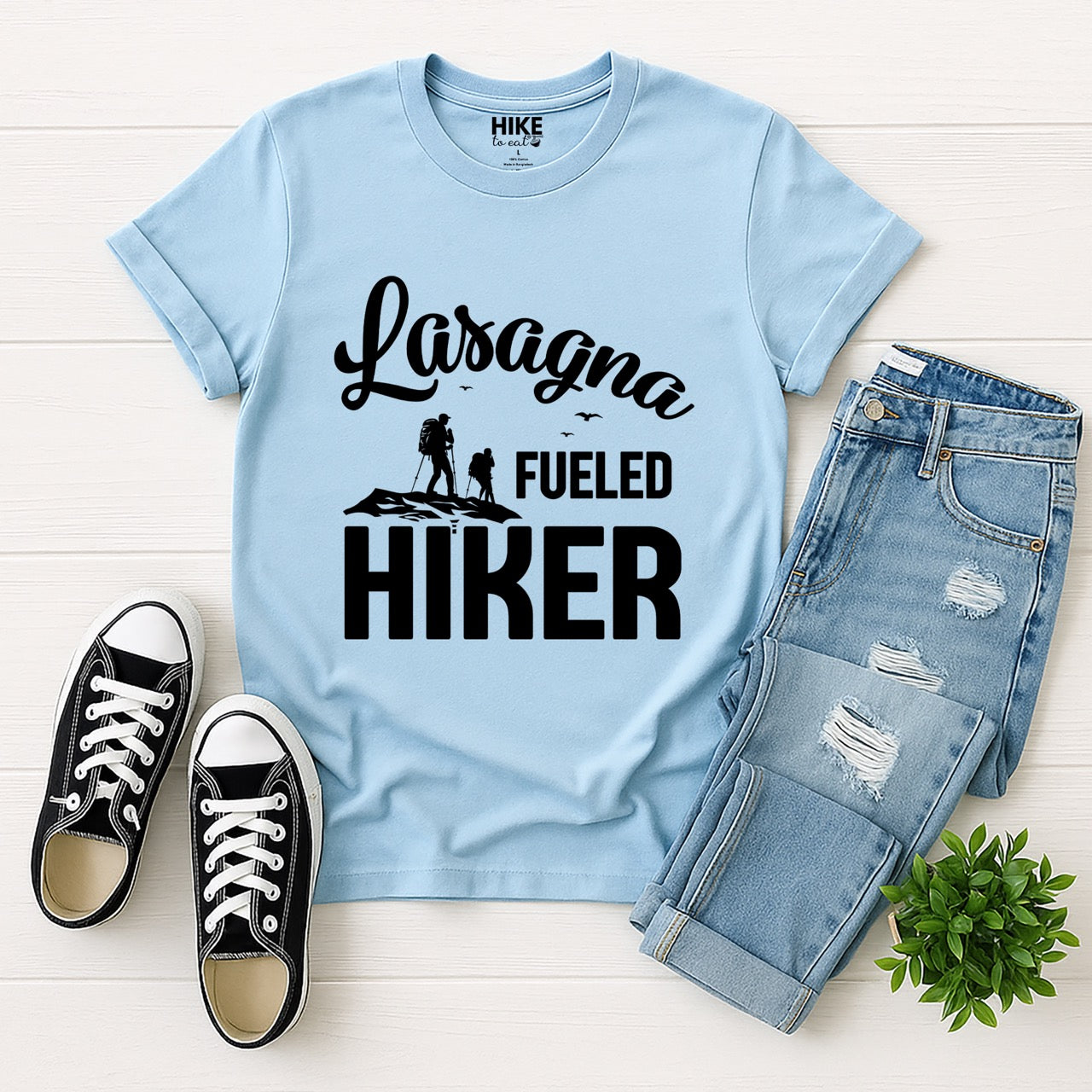 Light Blue Lasagna Fueled Hiker statement tee with flowing script typography, bold lettering, two hiker silhouettes on mountain ridge, and flying birds for Italian food-loving outdoor enthusiasts, hikers and foodies laid flat with shoes and pants
