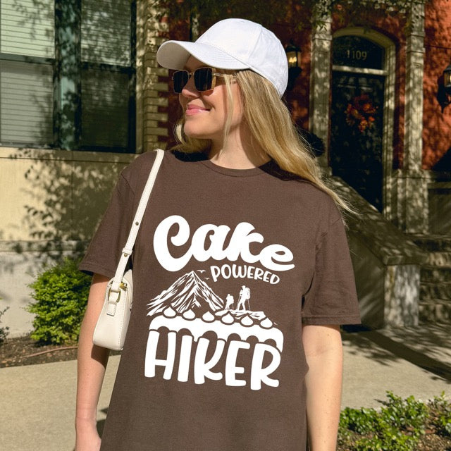 Woman wearing Dark chocolate Cake Powered Hiker statement tee with flowing script typography, bold lettering, mountain peaks, two hiker silhouettes, flying birds, and decorative cake illustration for dessert-loving outdoor enthusiasts, hikers and foodies