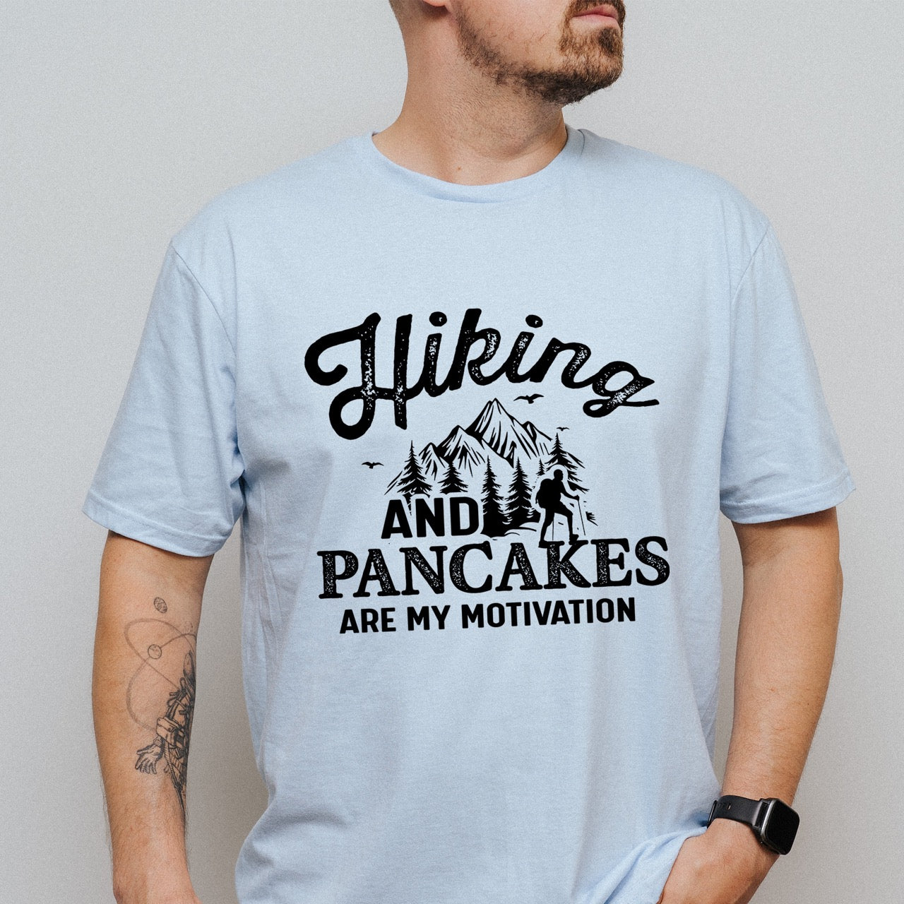 A man wearing Light Blue Hiking and Pancakes are my motivation statement tee with flowing script typography, bold lettering, mountain peaks, pine trees, hiker silhouette, and flying birds for pancake-loving outdoor enthusiasts, hikers and foodies