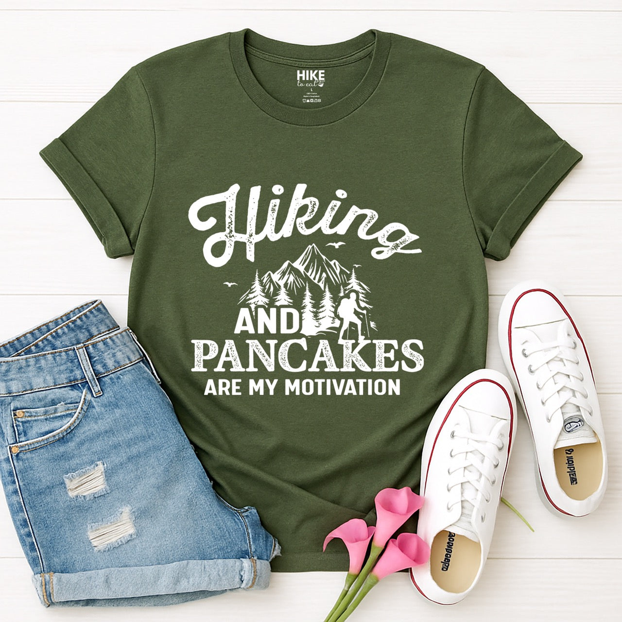 Military Green Hiking and Pancakes are my motivation statement tee with flowing script typography, bold lettering, mountain peaks, pine trees, hiker silhouette, and flying birds for pancake-loving outdoor enthusiasts, hikers and foodies laid flat with shorts and shoes