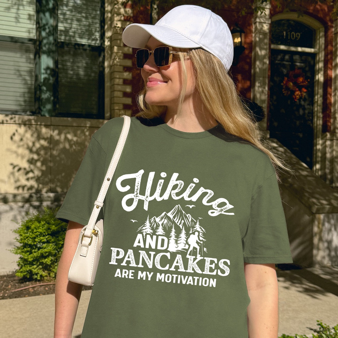 Woman wearing Military Green Hiking and Pancakes are my motivation statement tee with flowing script typography, bold lettering, mountain peaks, pine trees, hiker silhouette, and flying birds for pancake-loving outdoor enthusiasts, hikers and foodies