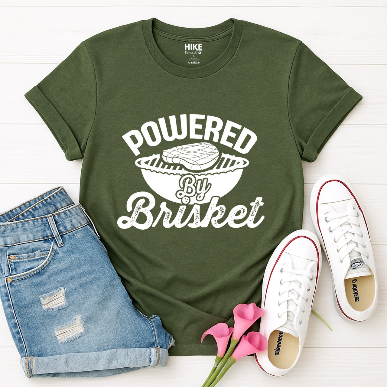 Military Green Powered by Brisket statement tee with bold curved typography, flowing script lettering, and BBQ grill with brisket illustration for barbecue-loving outdoor enthusiasts, hikers and foodies laid out with shorts and shoes