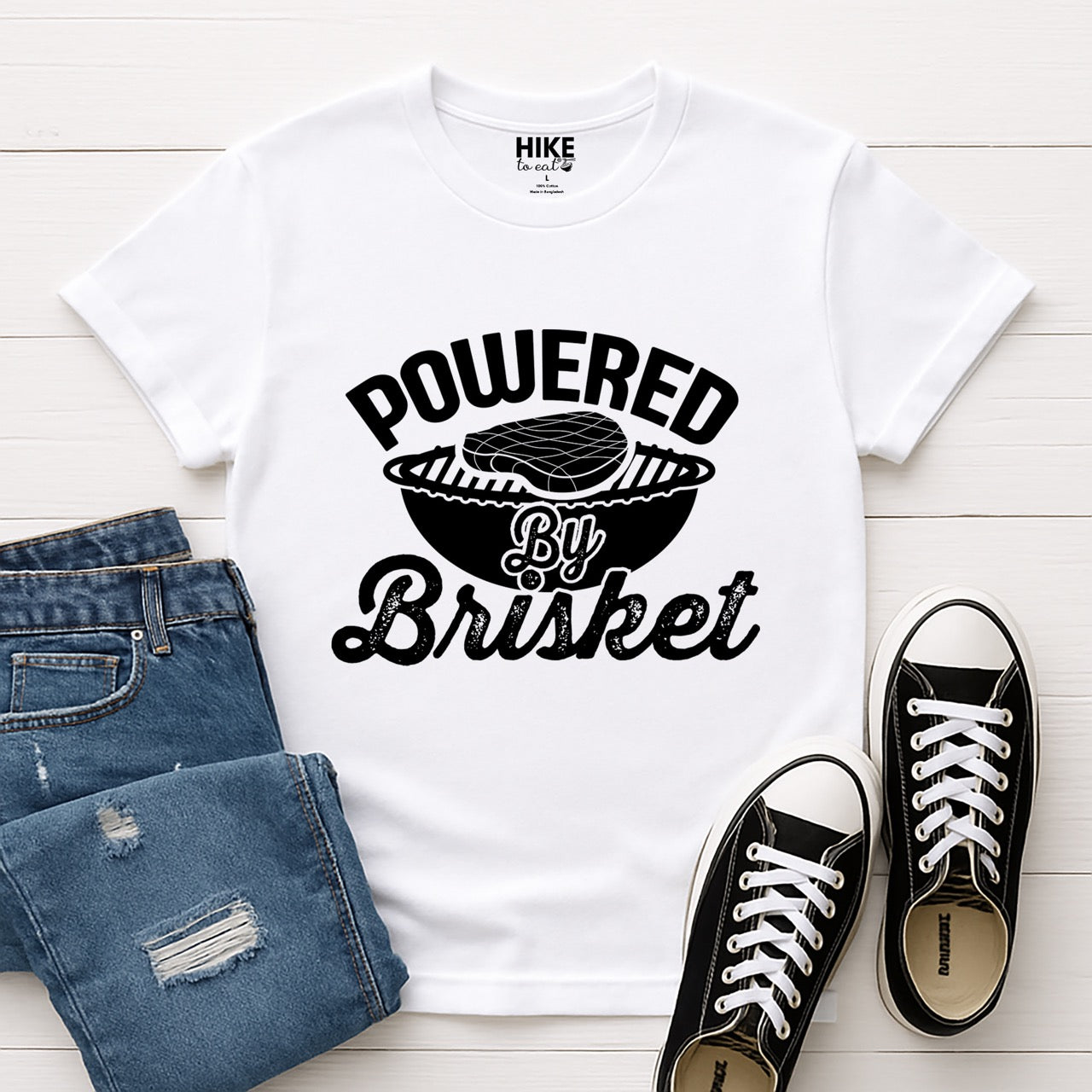 White Powered by Brisket statement tee with bold curved typography, flowing script lettering, and BBQ grill with brisket illustration for barbecue-loving outdoor enthusiasts, hikers and foodies laid flat with pants and shoes