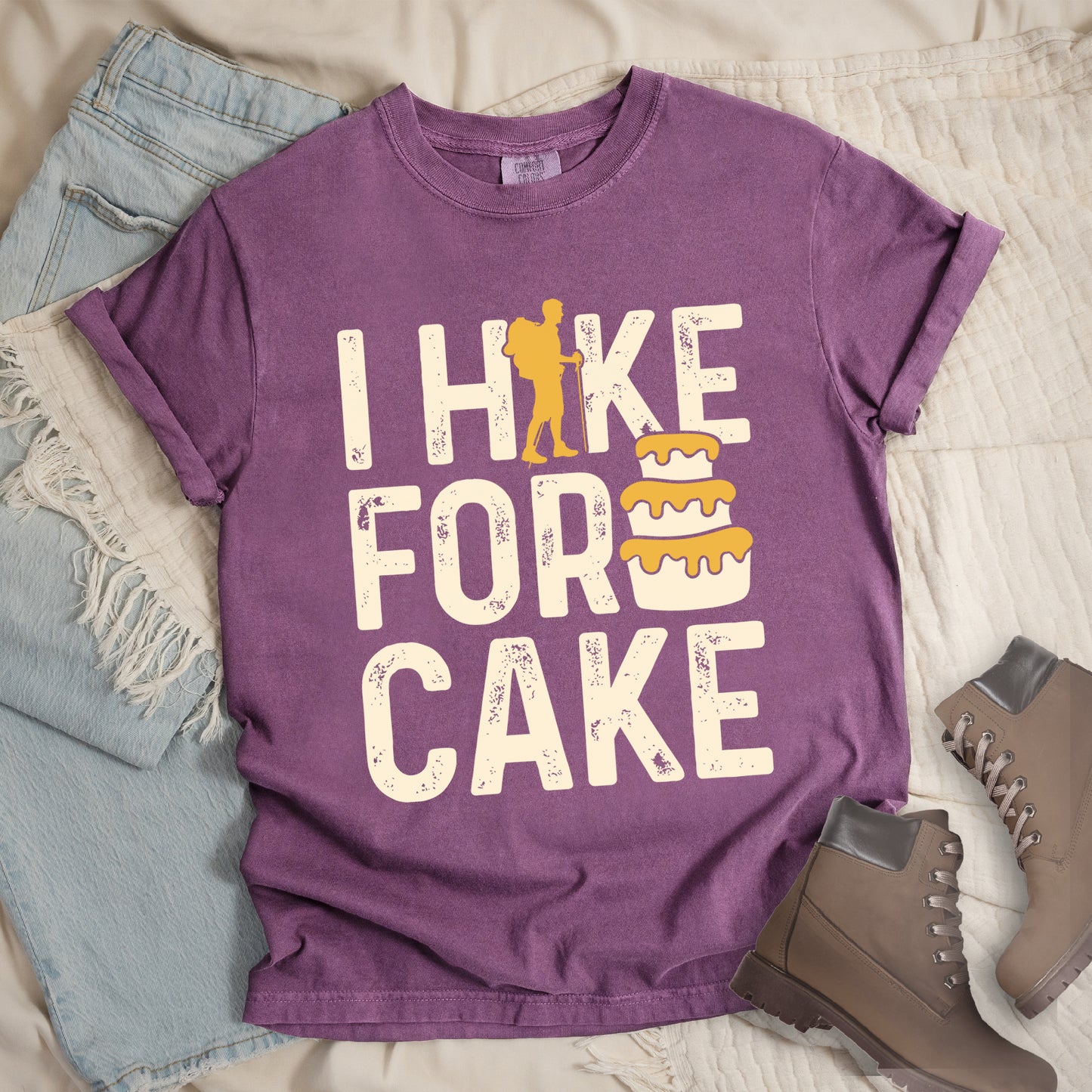 Berry (Dark Violet) t-shirt with distressed cream-colored text reading "I HIKE FOR CAKE". A yellow hiker silhouette is cleverly incorporated into the letter "K," while a three-tiered cake with dripping yellow icing appears between "FOR" and "CAKE." The shirt features a classic crew neck and casual rolled sleeves, styled alongside light wash denim and brown hiking boots.
