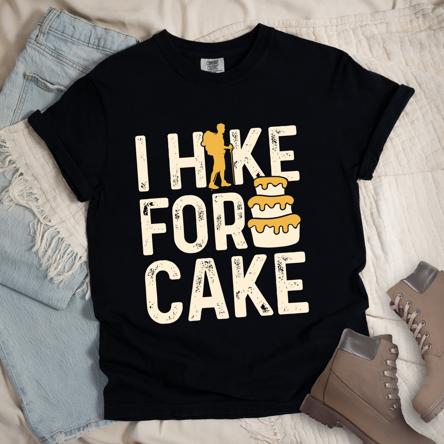 The black t-shirt displays "I HIKE FOR CAKE" in distressed off-white lettering. A yellow hiker silhouette is cleverly incorporated into the letter "K," while a three-tiered cake with dripping yellow icing appears between "FOR" and "CAKE." The shirt features a classic crew neck and casual rolled sleeves, styled alongside light wash denim and brown hiking boots.